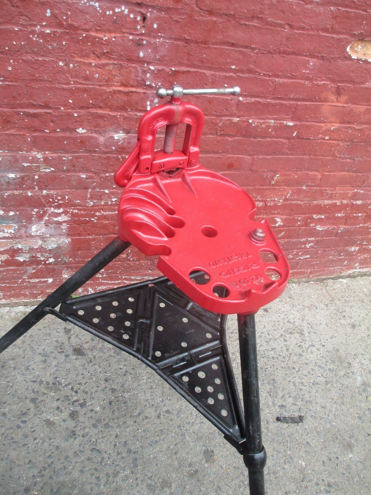 A red pipe clamp is sitting on a tripod in front of a brick wall.