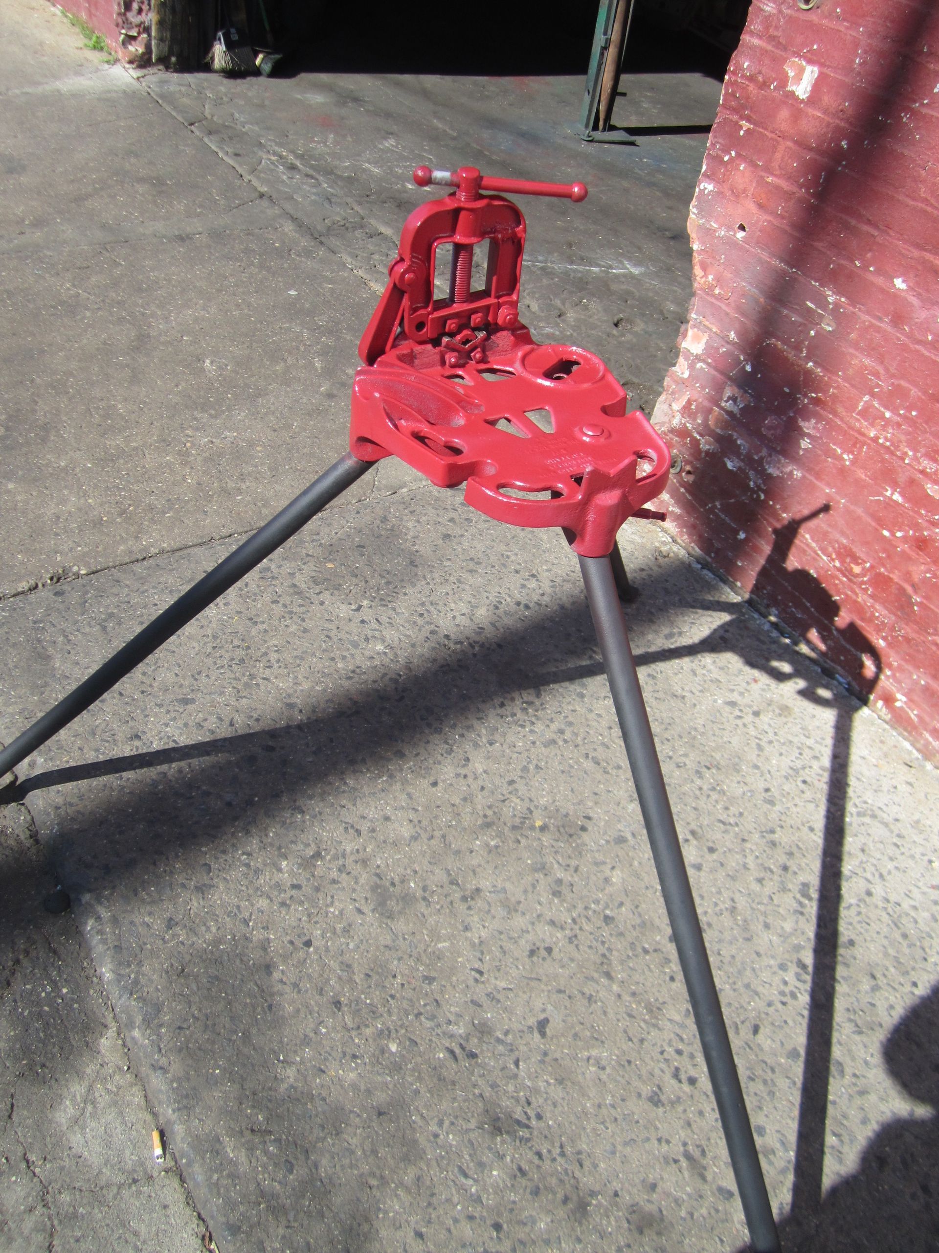 A red pipe vise is sitting on the sidewalk in front of a brick wall