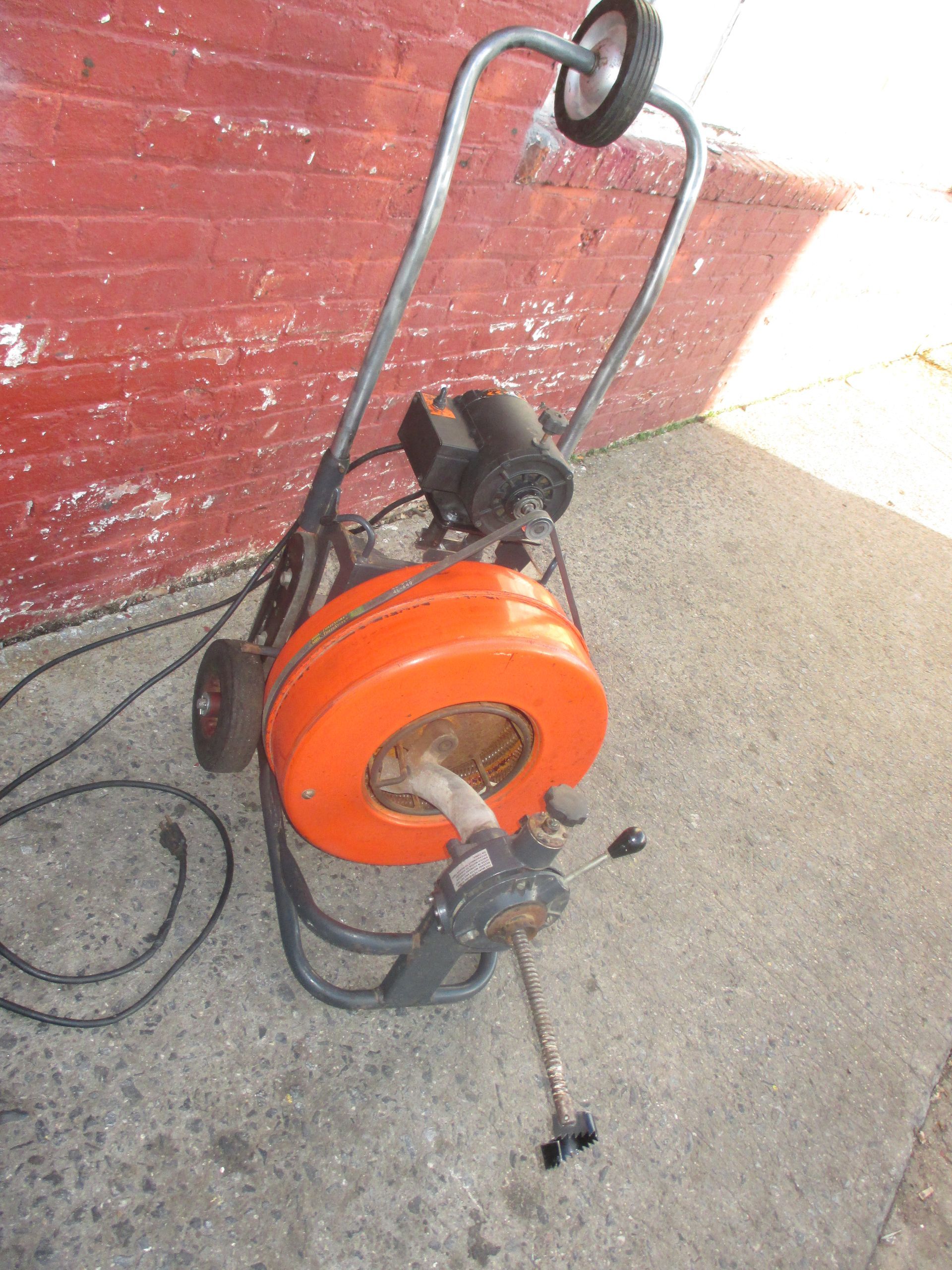 A drain cleaner is sitting on the ground in front of a brick wall