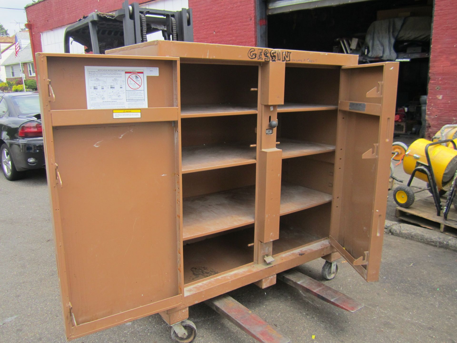 A brown cabinet with the doors open is sitting on a forklift in front of a garage.