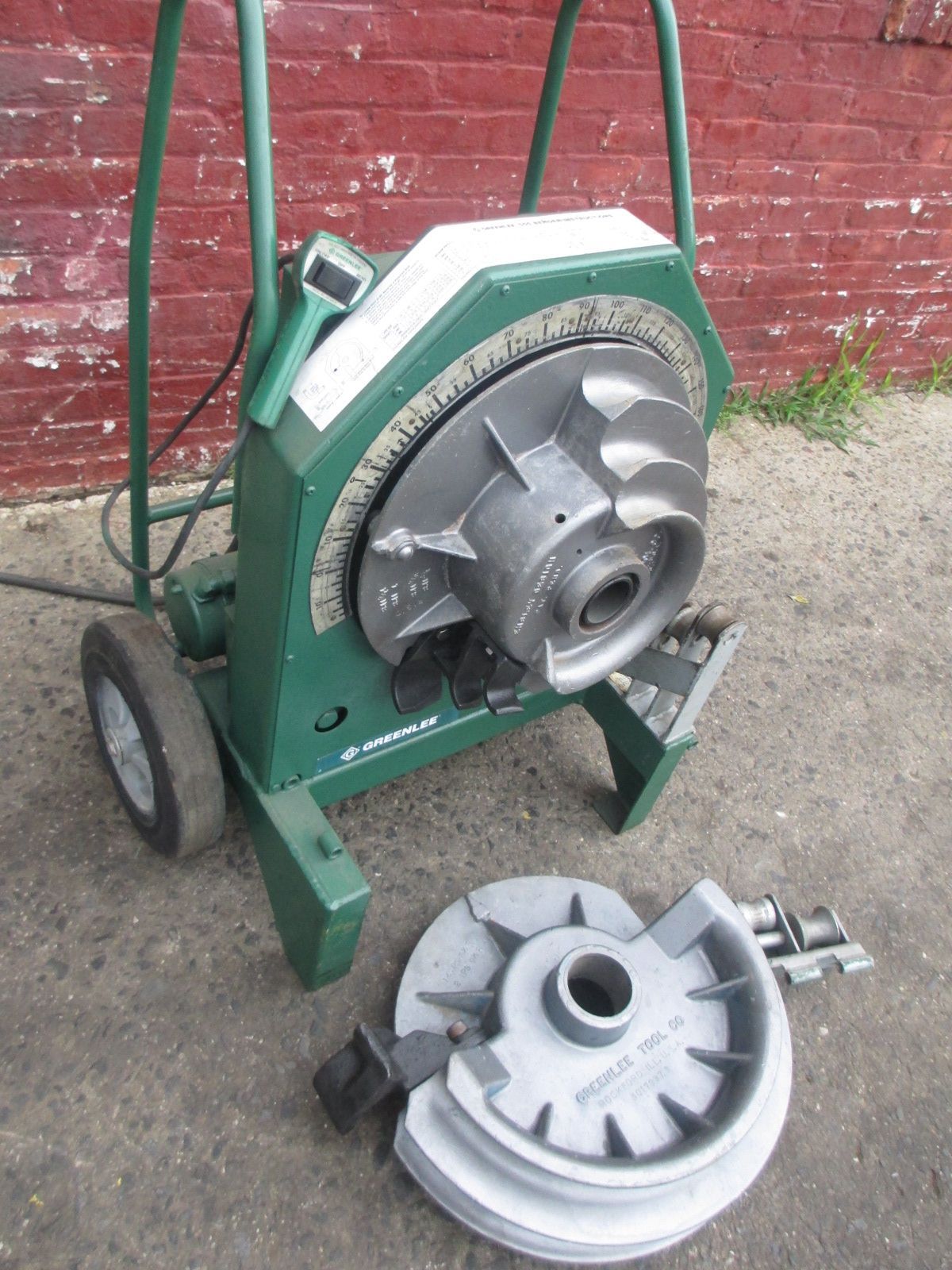 A green pipe bending machine is sitting in front of a red brick wall
