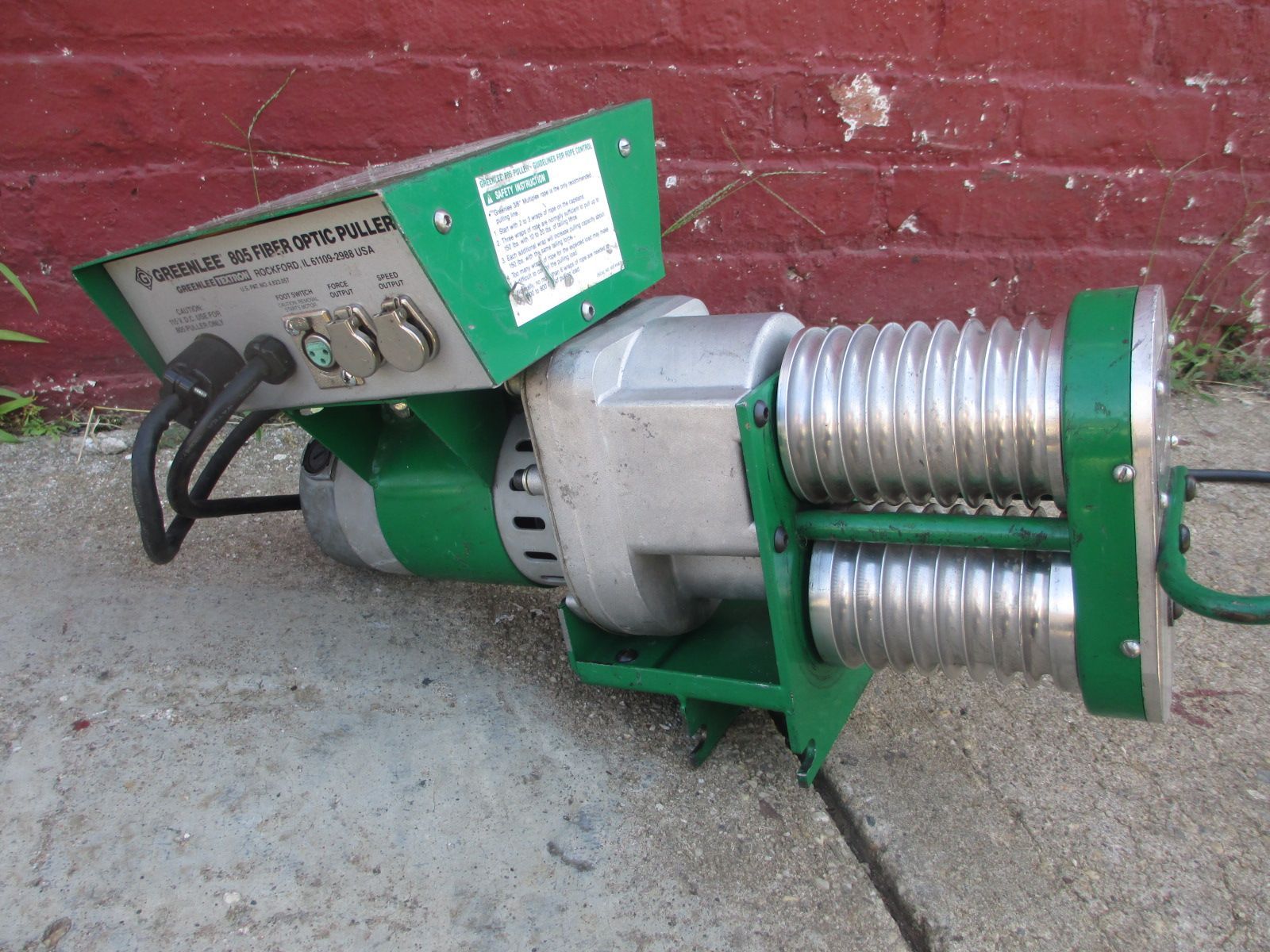 A green machine is sitting on a sidewalk in front of a red brick wall
