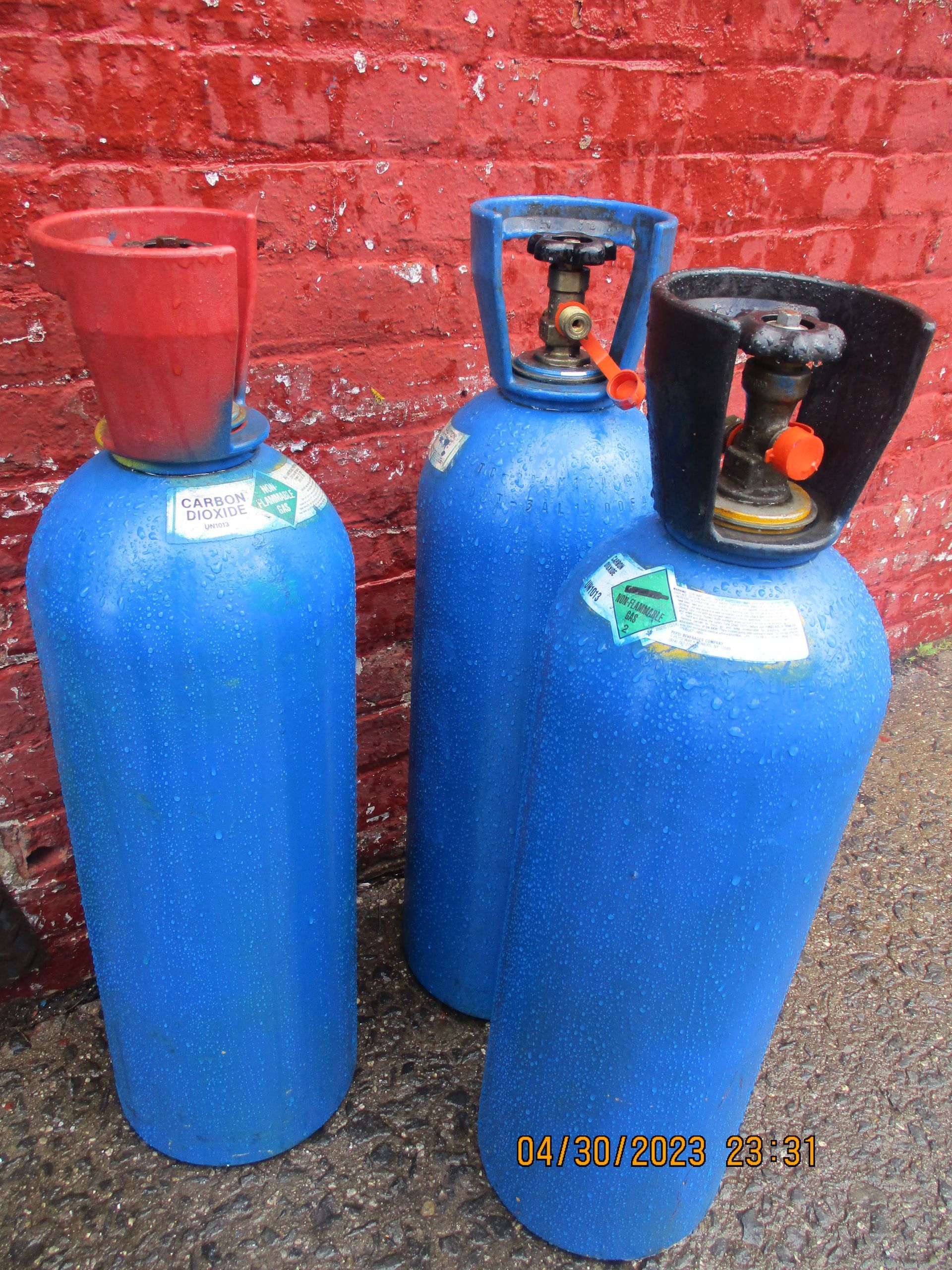 Three blue gas cylinders are sitting in front of a red brick wall