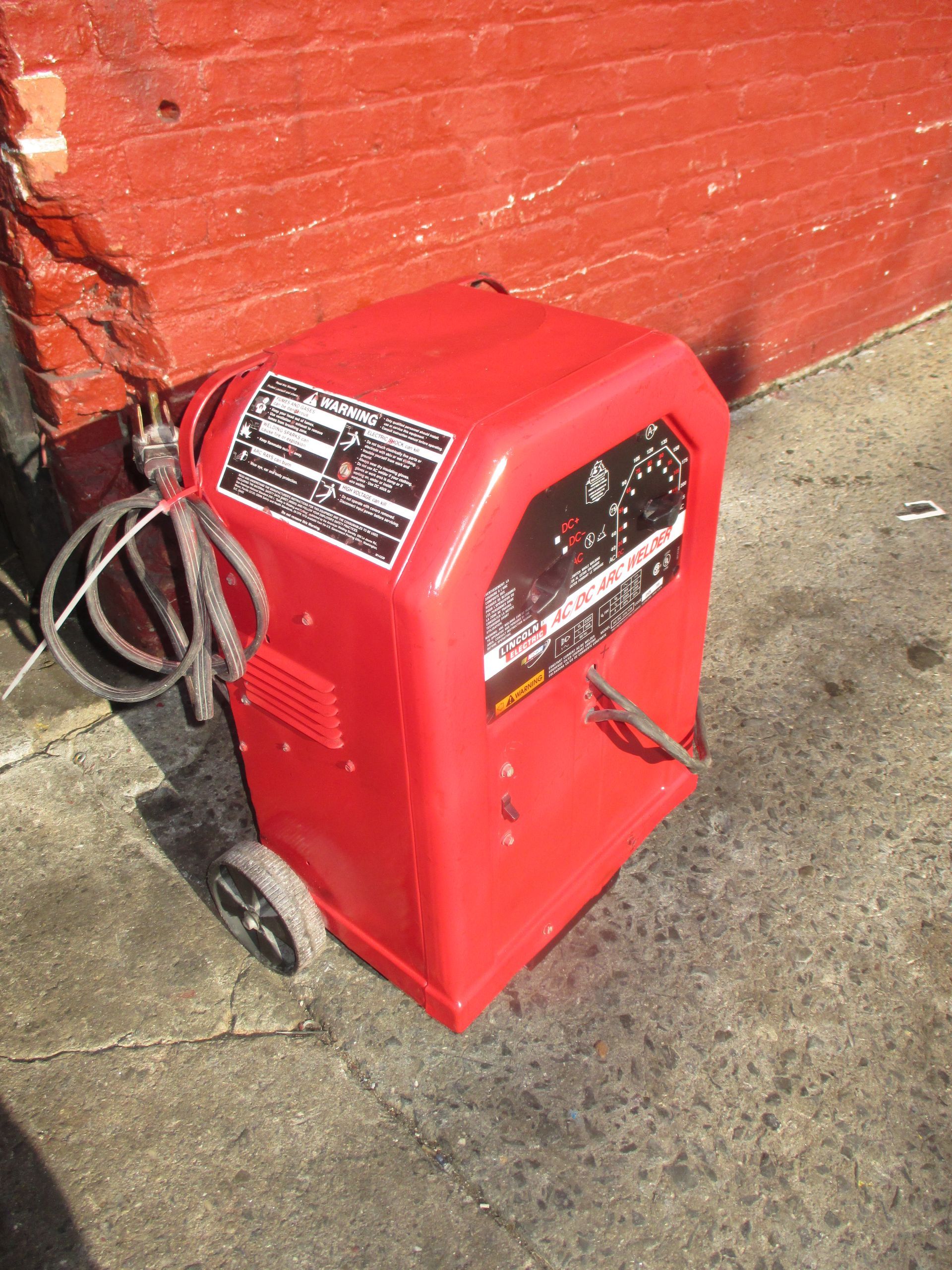 A red welding machine is sitting on the sidewalk in front of a brick wall.