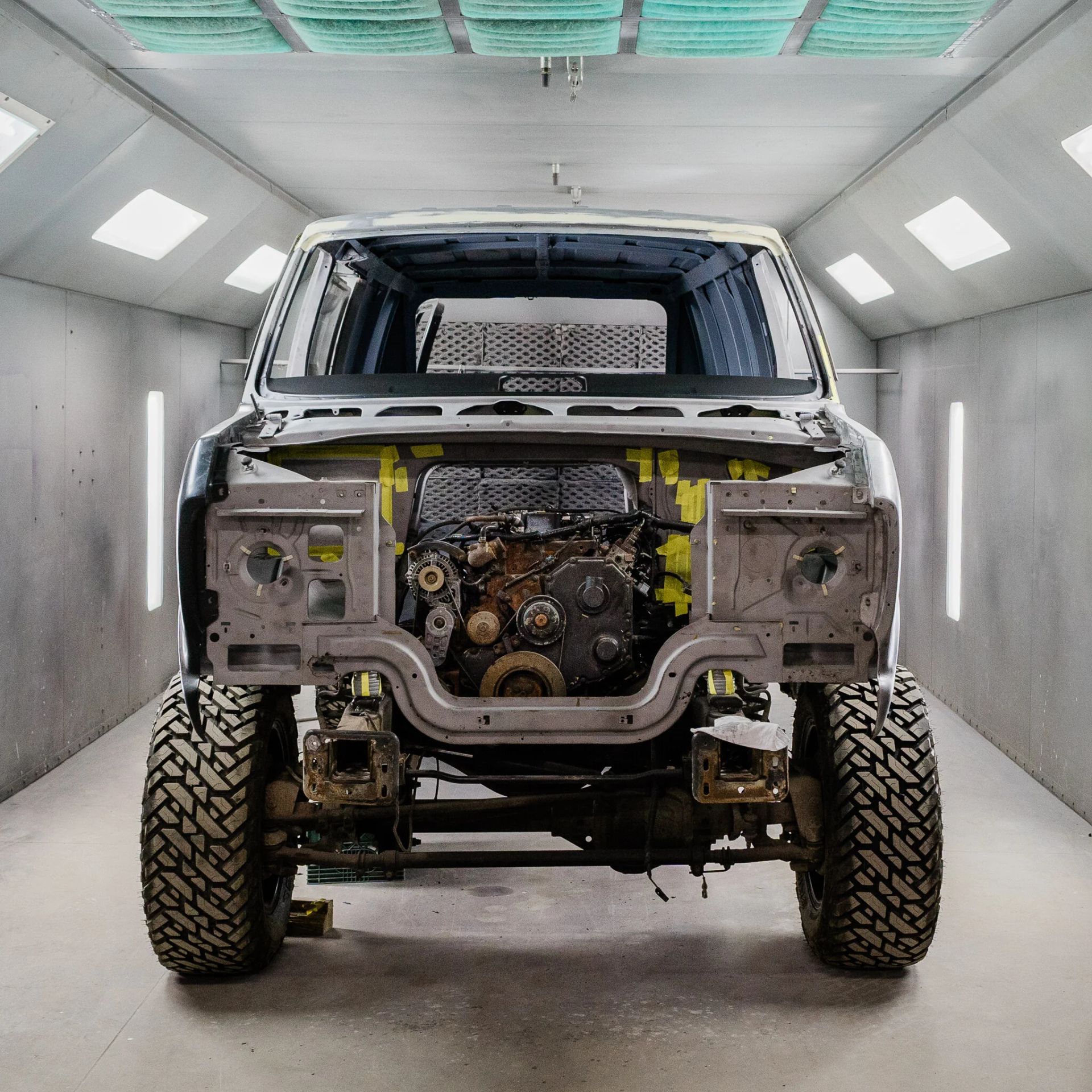 Front view of a rugged off-road vehicle inside a white spray booth, with its grille and front body panels removed.