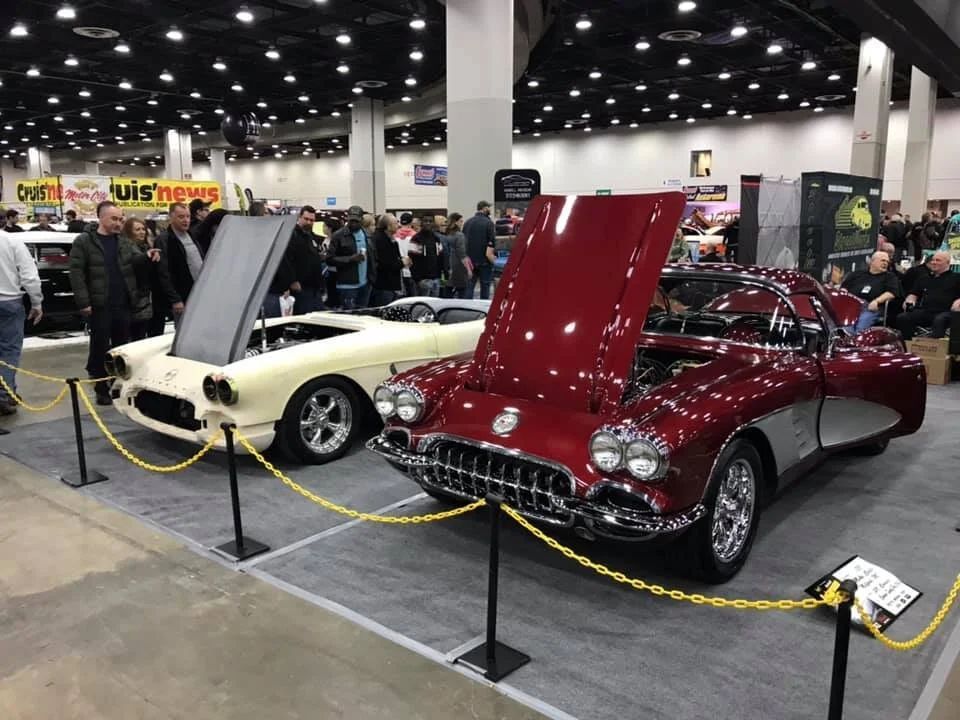 Two classic cars on display at an indoor auto show, one cream and one red, both with hoods open