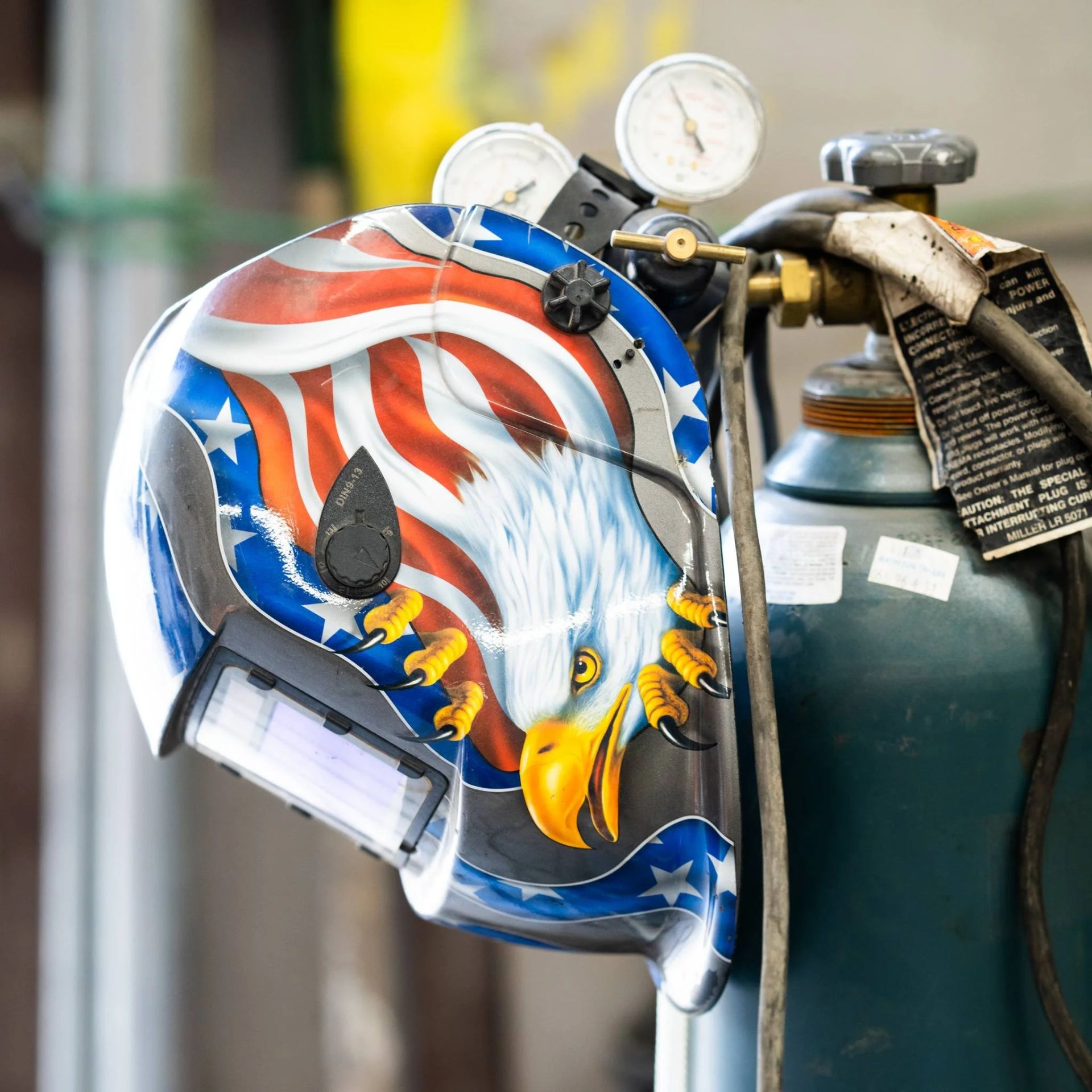 Painted welding helmet with an eagle and U.S. flag design hanging on a gas cylinder
