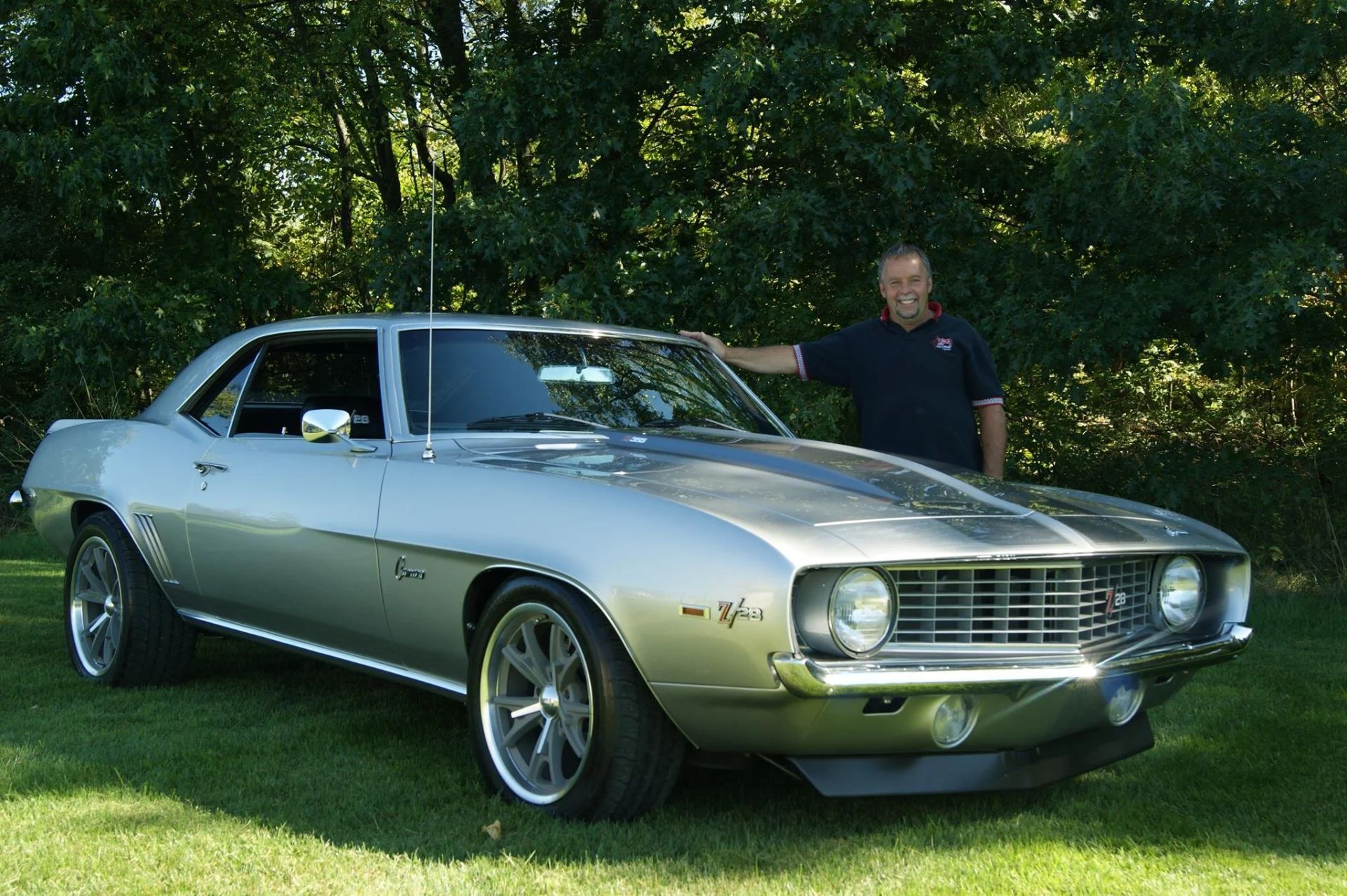 Silver classic muscle car parked on grass with a man standing behind it in a wooded area