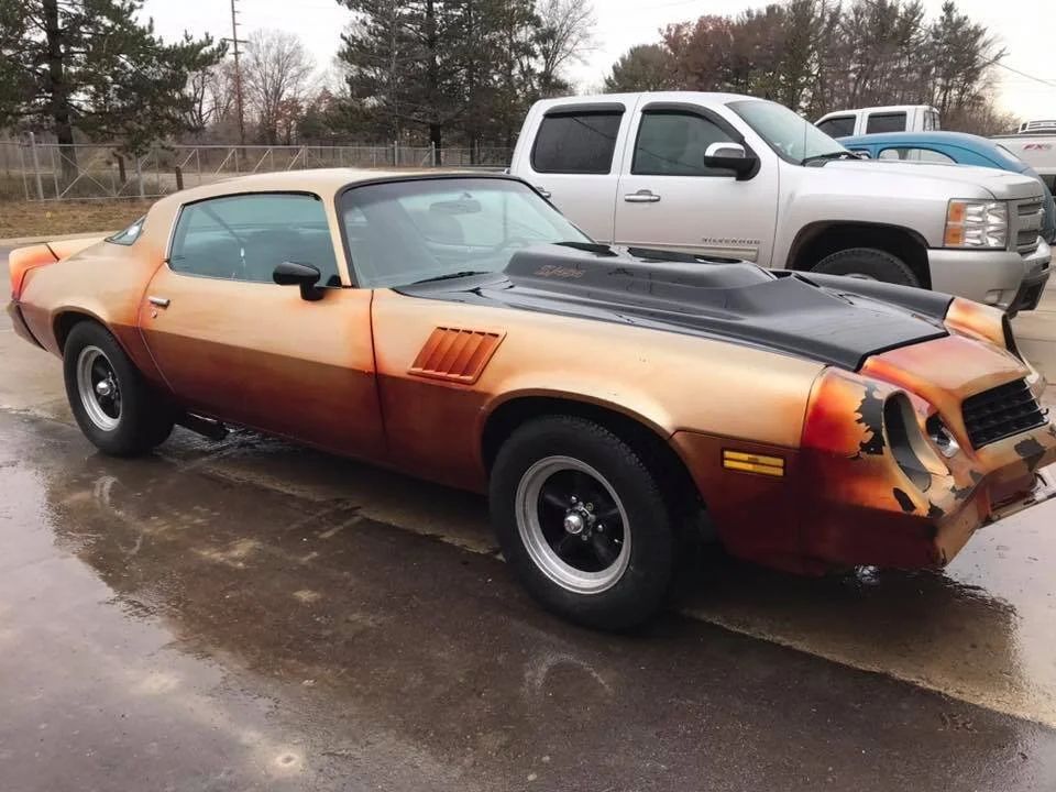 Old orange muscle car with front-end damage parked in a wet lot, with a white pickup behind it