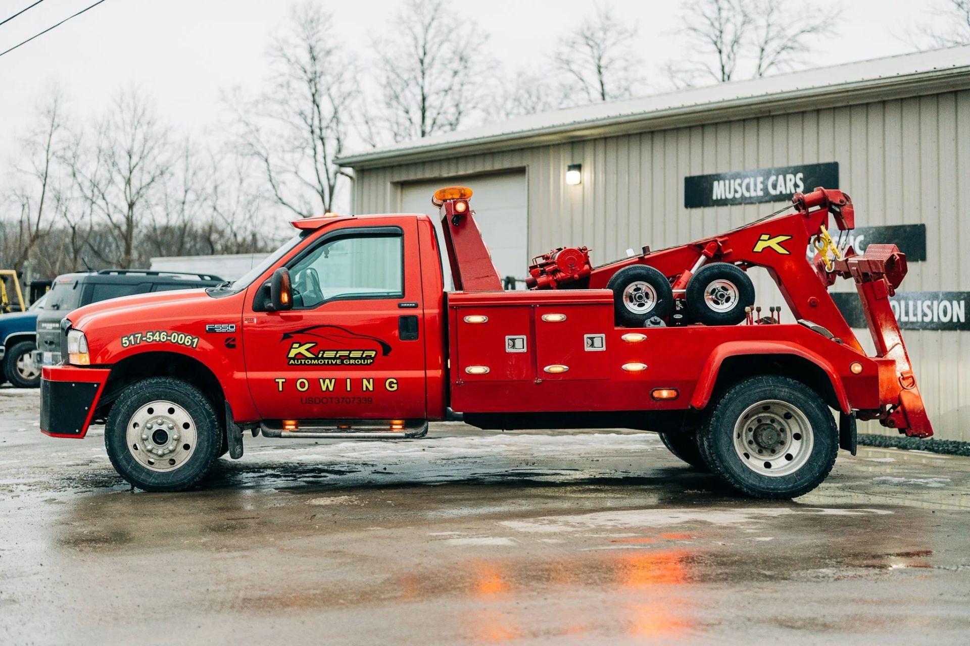 Red tow truck parked outside a building on a wet road, with bare trees in the background.