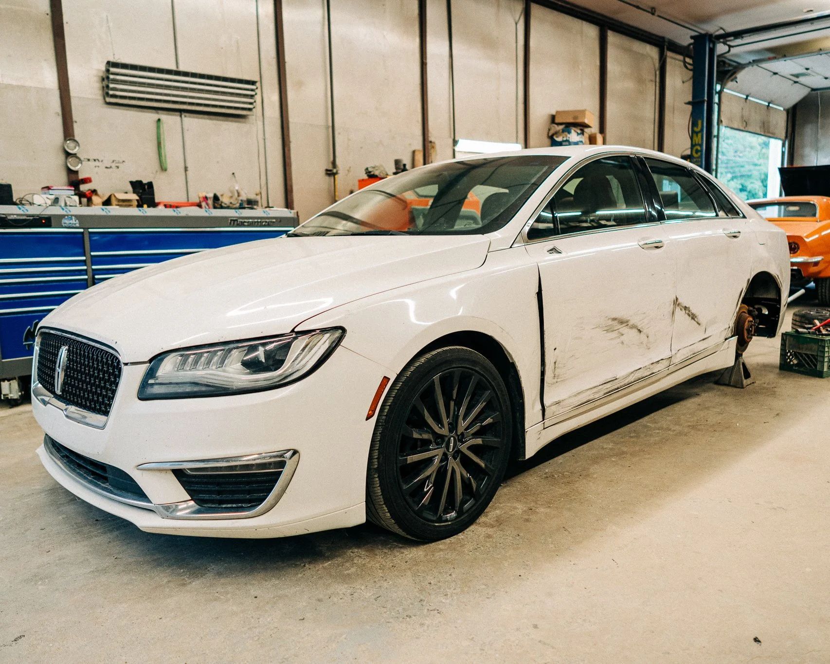 White luxury sedan in an auto shop with minor side damage and a removed rear wheel