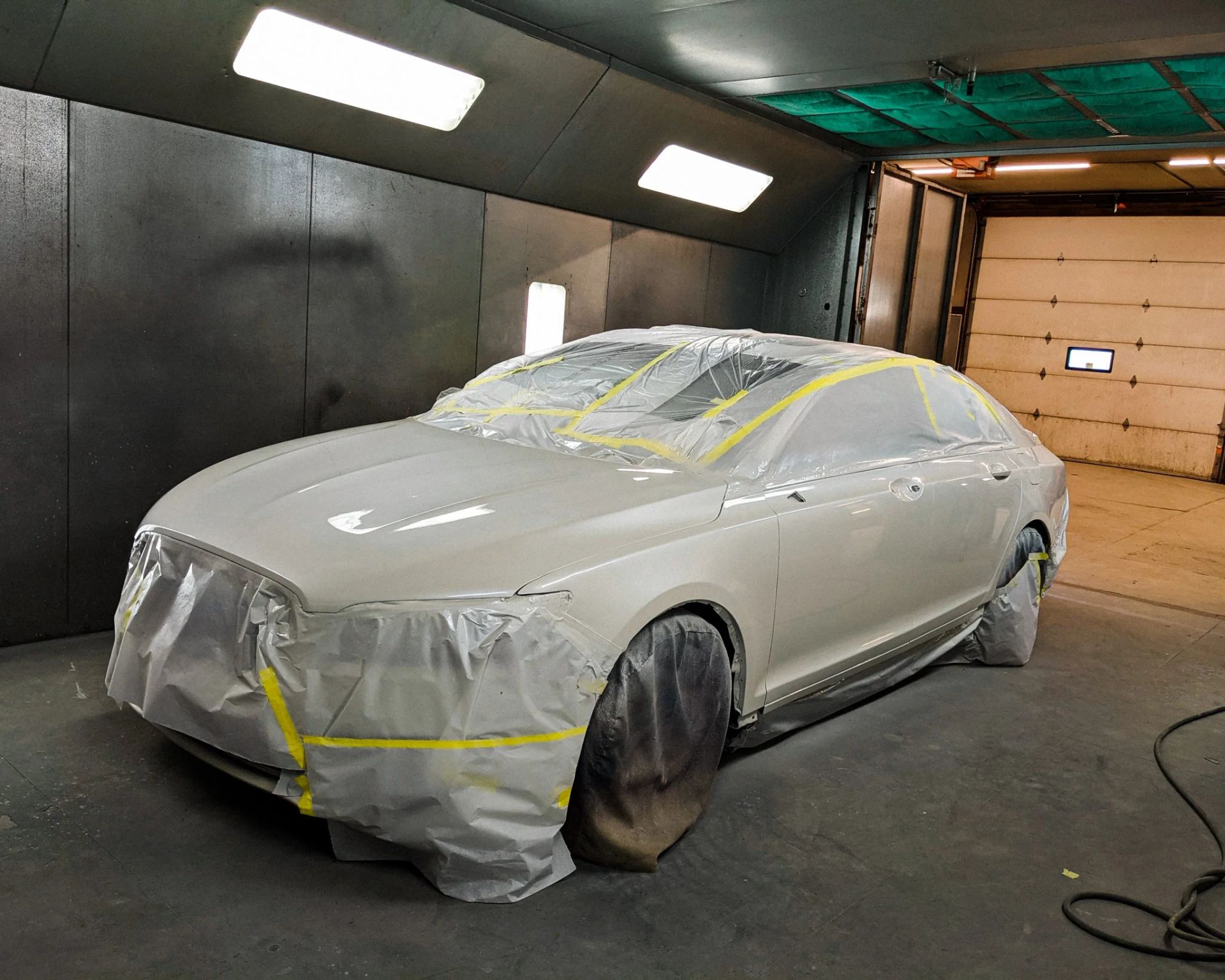 Silver car covered in masking tape and plastic in a spray booth, likely being painted