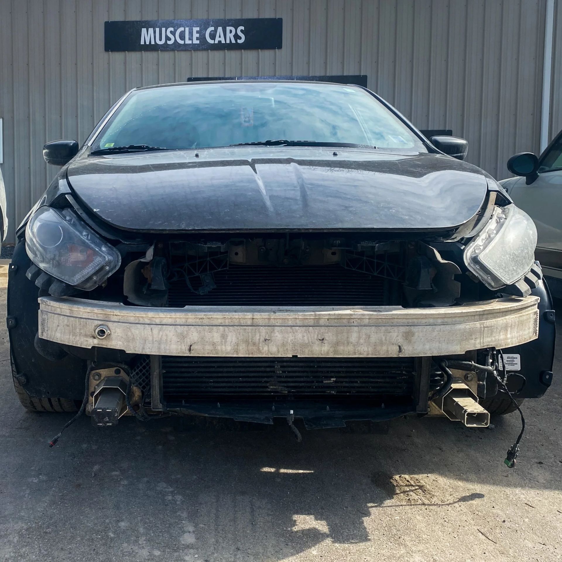 Front view of a damaged black car with a missing grille, parked outside a Muscle Cars shop.