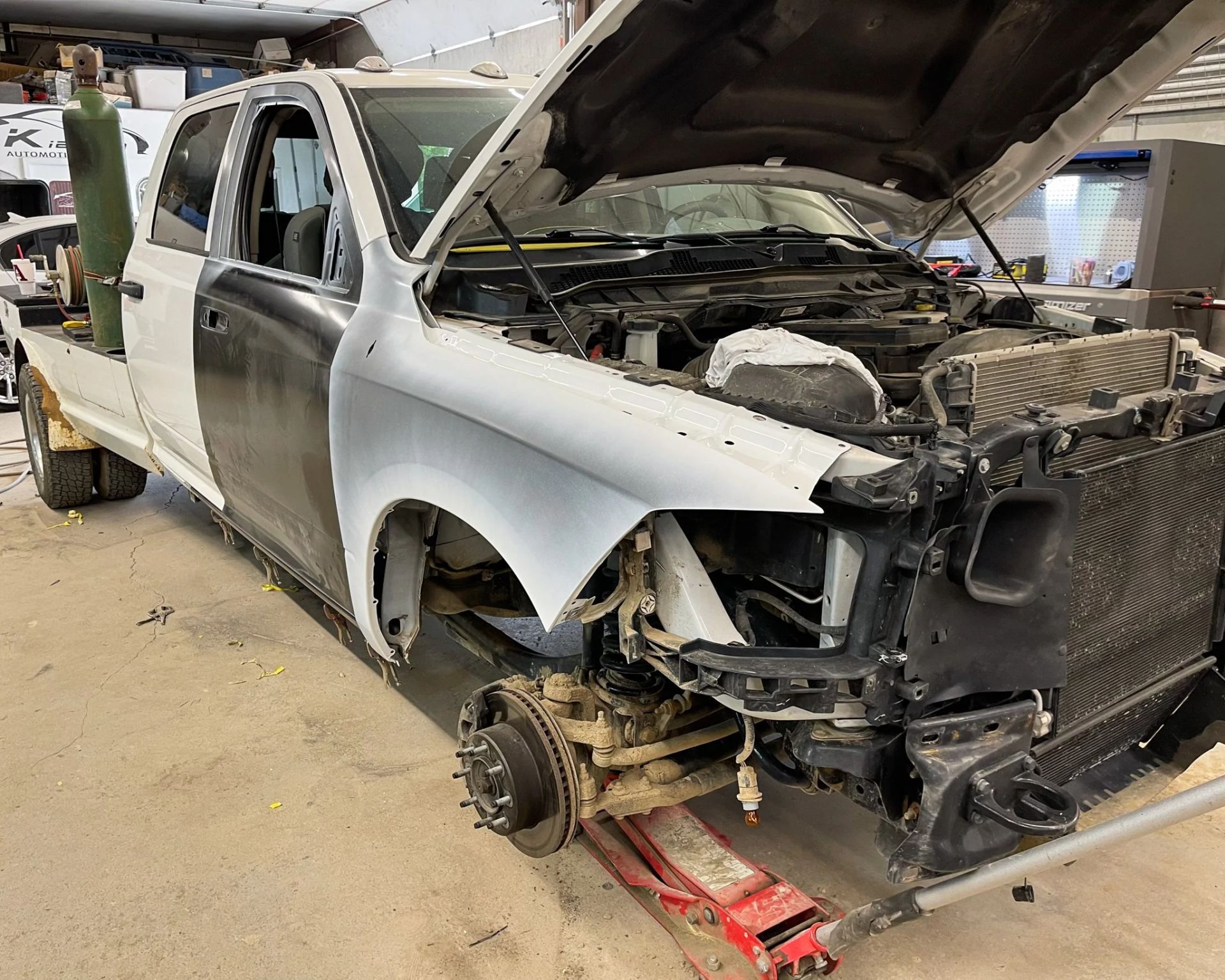 Damaged white pickup truck in a repair shop, front end torn apart and wheel removed on a lift.