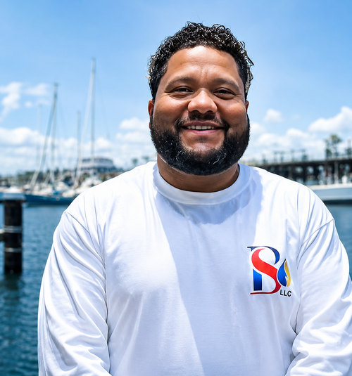 Smiling man in a white long-sleeve shirt standing by a marina with boats and blue sky.
