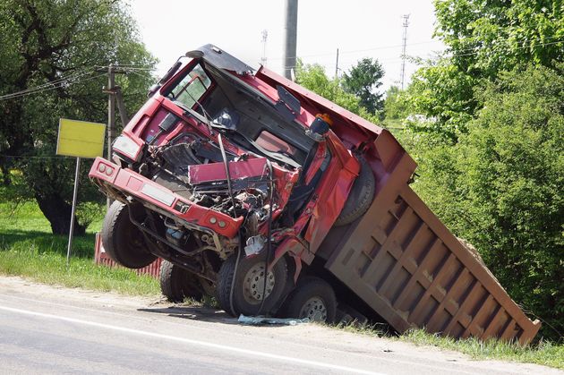 Red dump truck overturned in a roadside ditch, cab crushed and trailer tipped on its side.