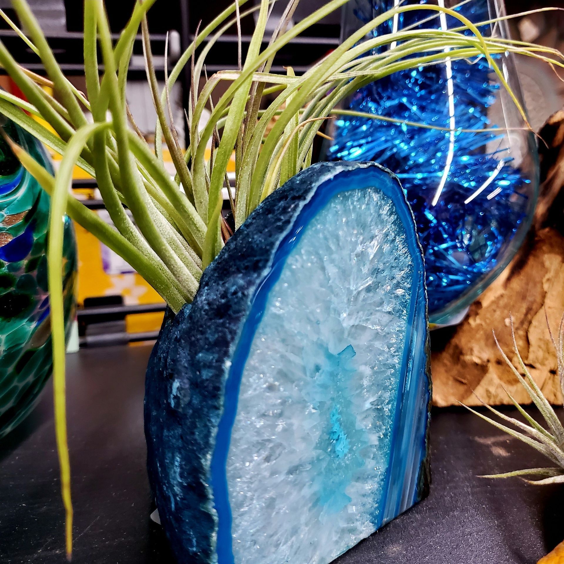 An air plant sits in a blue geode planter, positioned next to a glass bowl filled with blue decorative shreds.