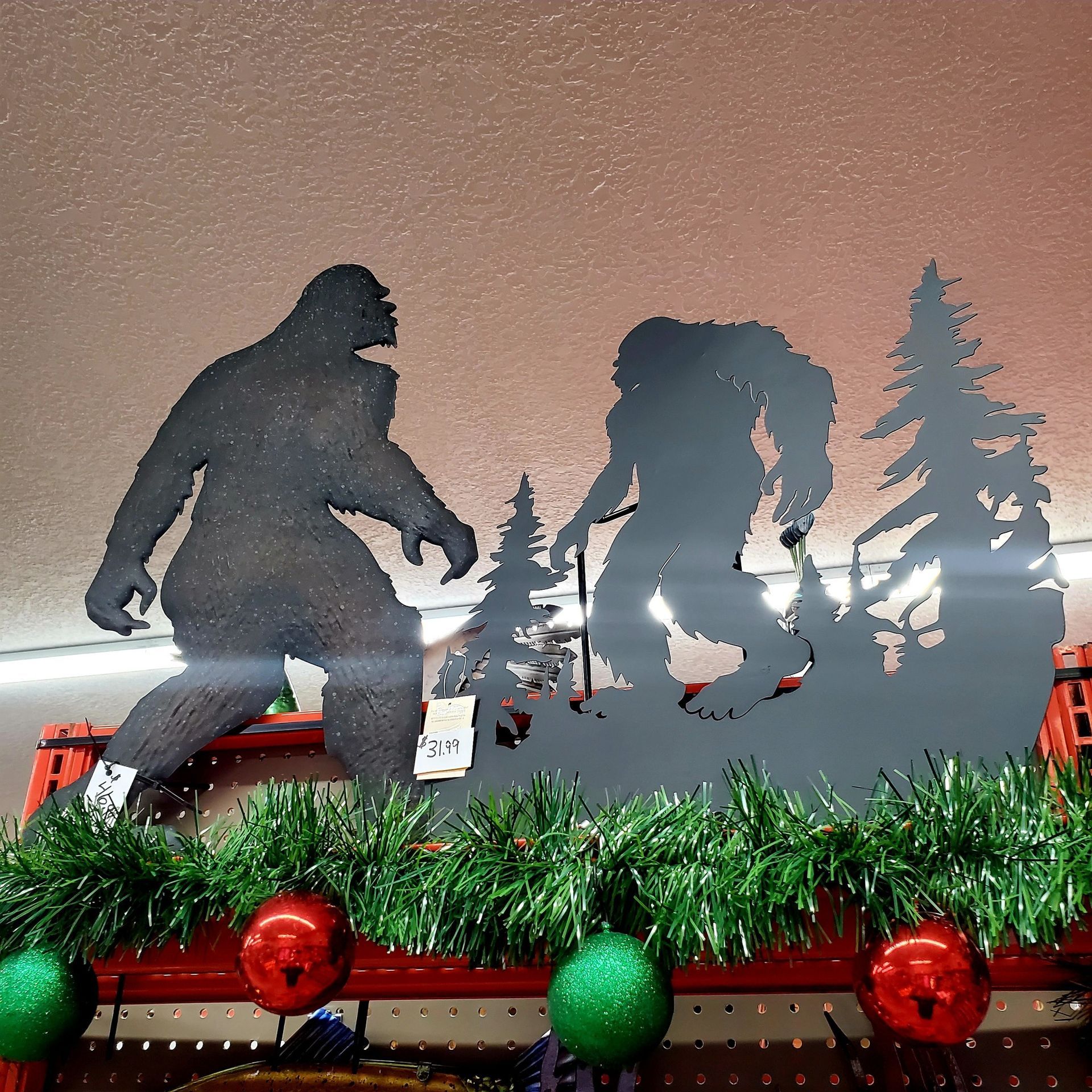 A metal wall art silhouette of two Sasquatches in a forest, displayed on a shelf with green garland and Christmas bulbs.