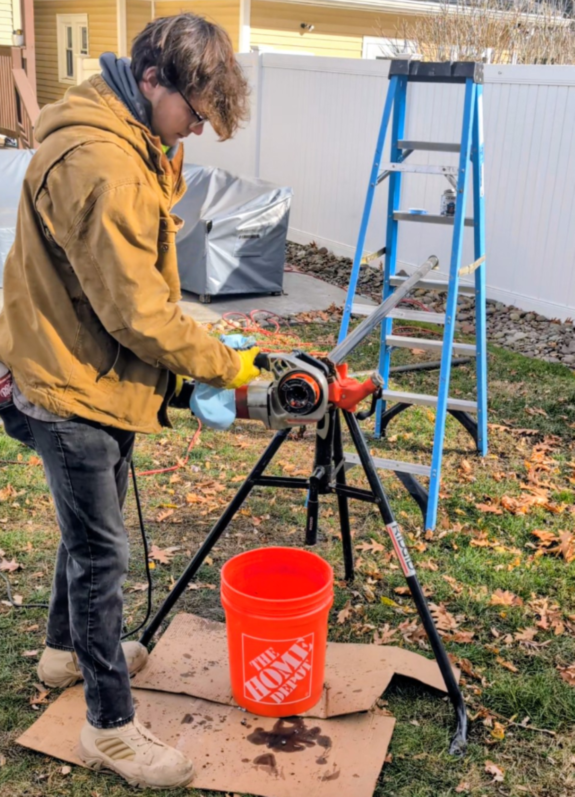 Man in a yellow jacket operates a pipe cleaner near a ladder in a yard, with a Home Depot bucket below.