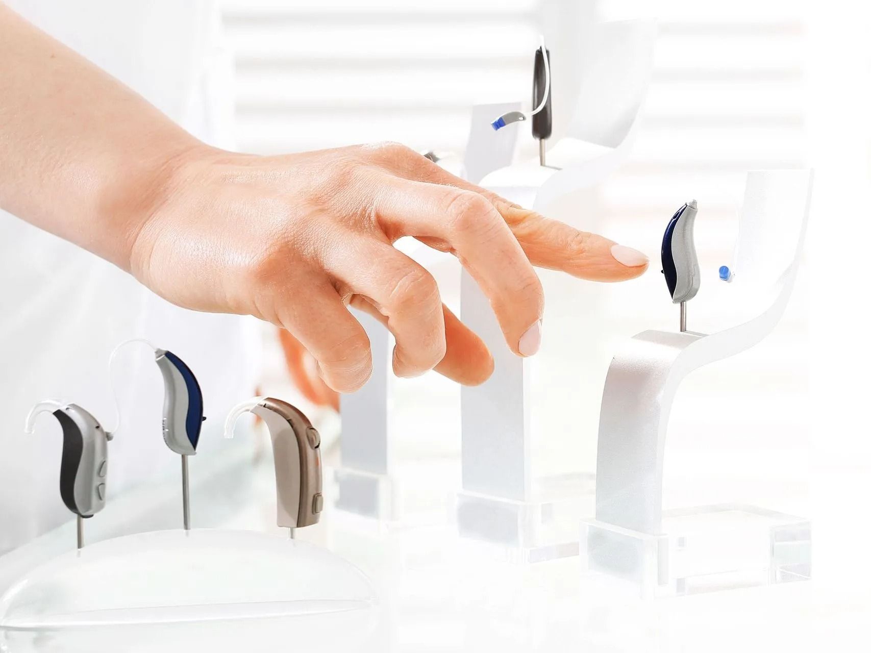 Hand pointing to a hearing aid on a white display, various models in view. Bright, clinical setting.