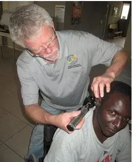 Man examining another man's ear with a tool; indoors. The man getting examined is wearing a gray shirt and looking towards the camera.