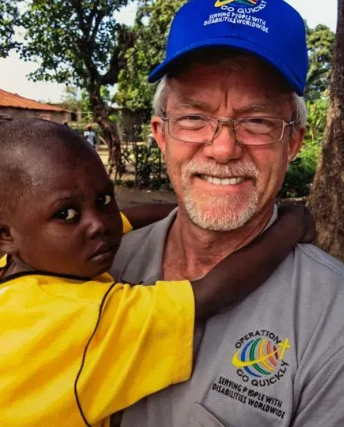 Man in blue cap holds child in yellow shirt. Both are smiling outdoors. Text on cap and shirt.