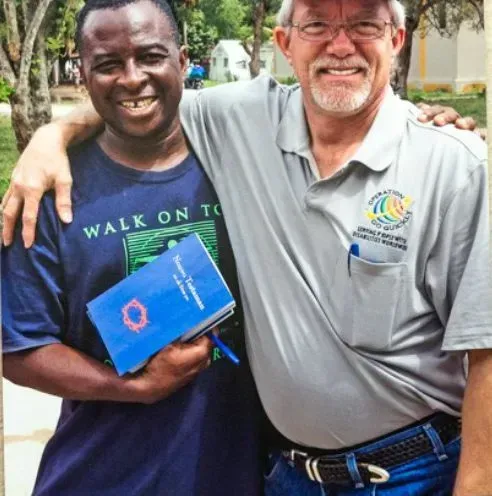 Two men, one holding a book, smile while embracing outdoors. One wears a grey shirt, the other a blue shirt.