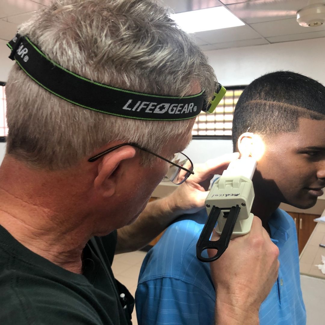 Man examining a patient's ear with a light. The patient smiles, indoors, bright light.