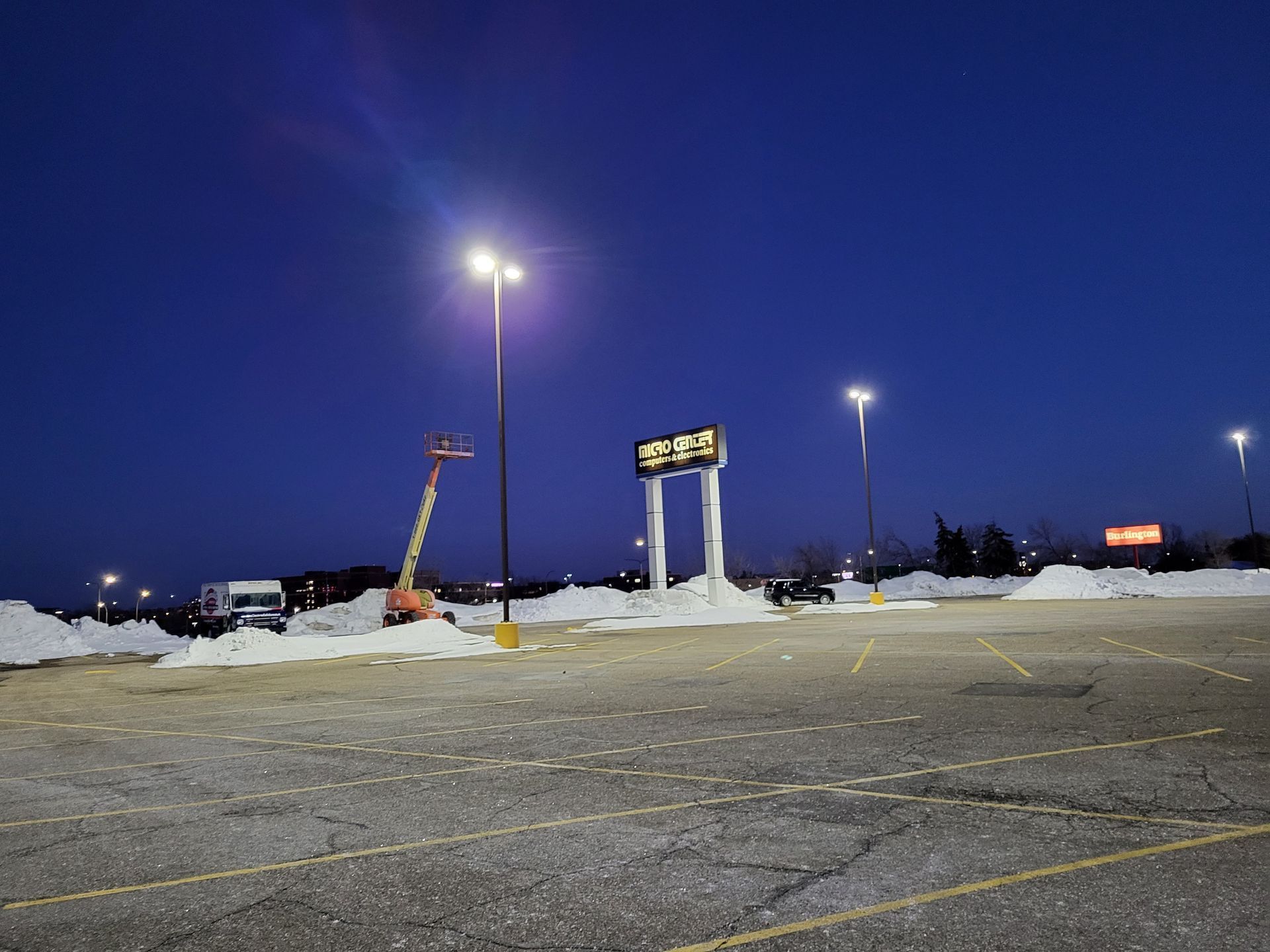 Empty parking lot at night with snow banks, under streetlights, and a lit sign.