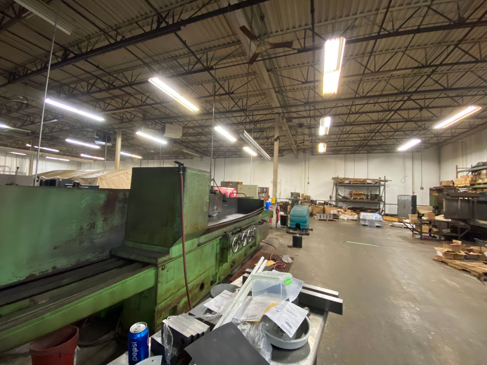 Inside a large industrial workshop with overhead lights, a green metal machine sits near a cluttered workspace.