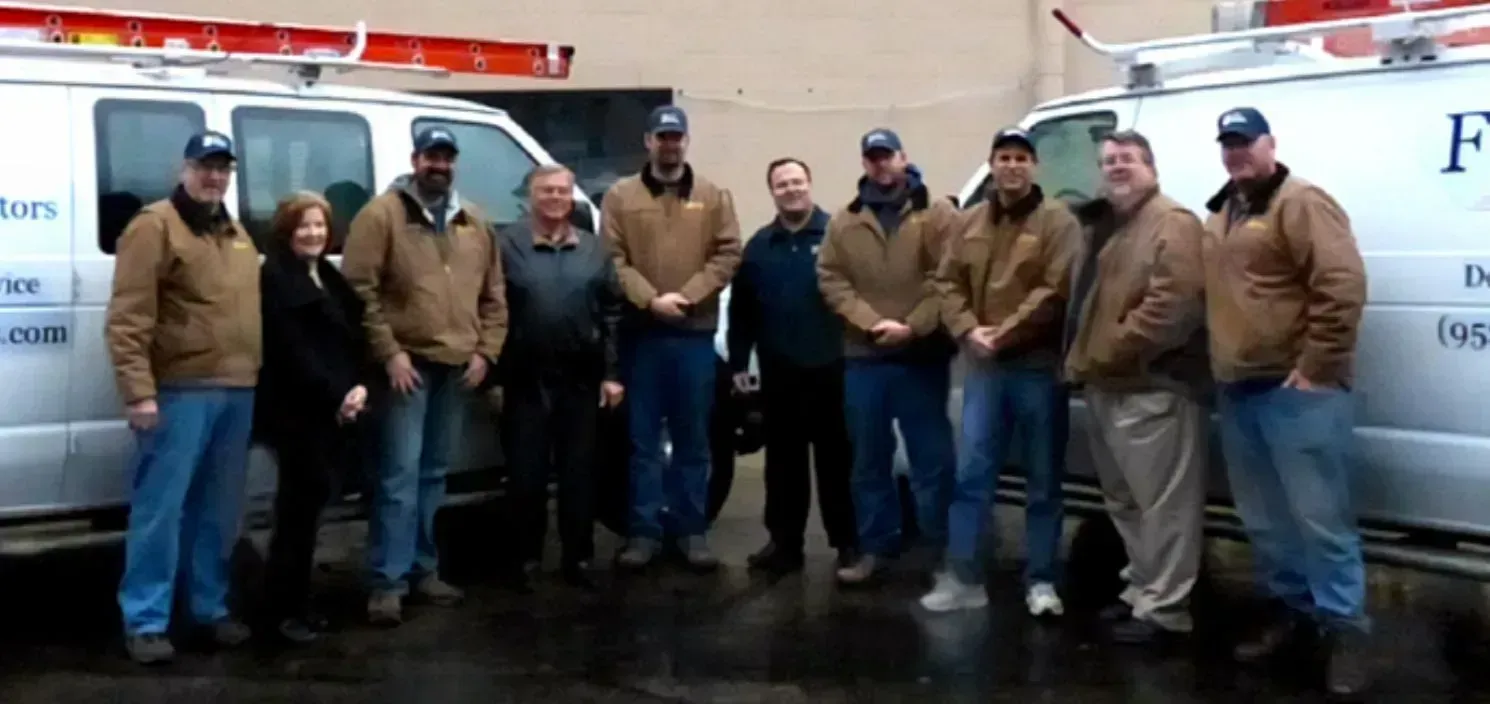 A group of people standing in front of two white vans. They are wearing brown jackets and blue jeans.