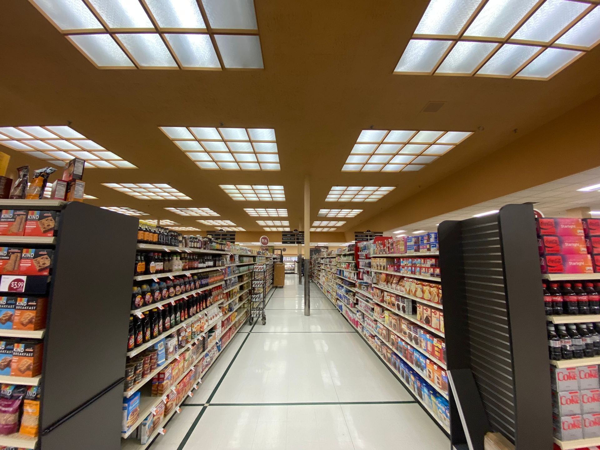 Grocery store aisle with shelves stocked with products, overhead fluorescent lights.