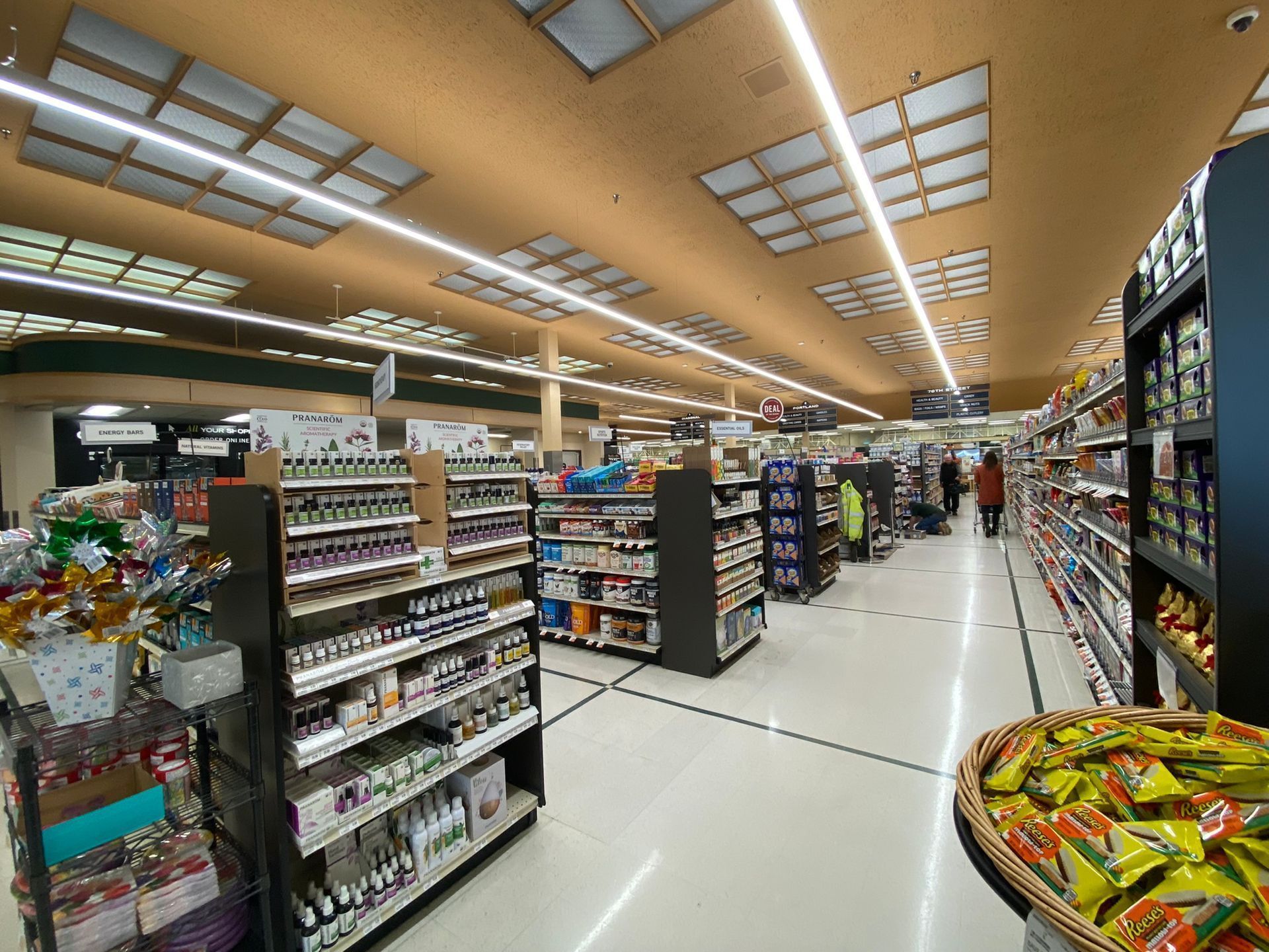Grocery store aisle with shelves of products and shoppers browsing. Overhead lighting.