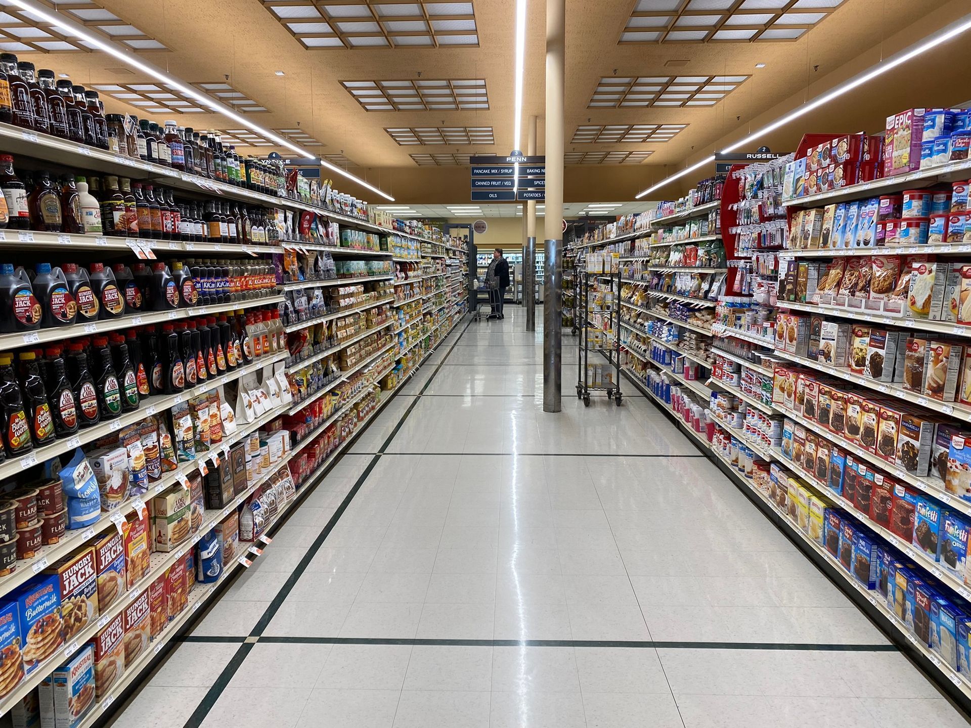 Grocery store aisle with shelves of food products, bright overhead lights, and a customer in the distance.