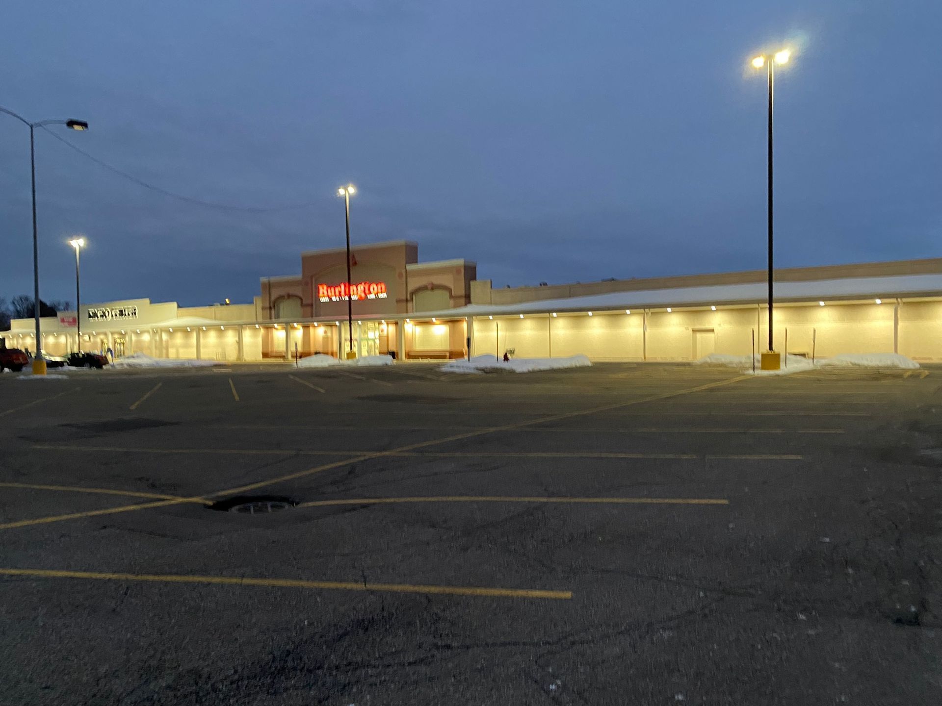 Empty parking lot at dusk, with snow on the edges. Lit sign and streetlights in a cold, overcast setting.