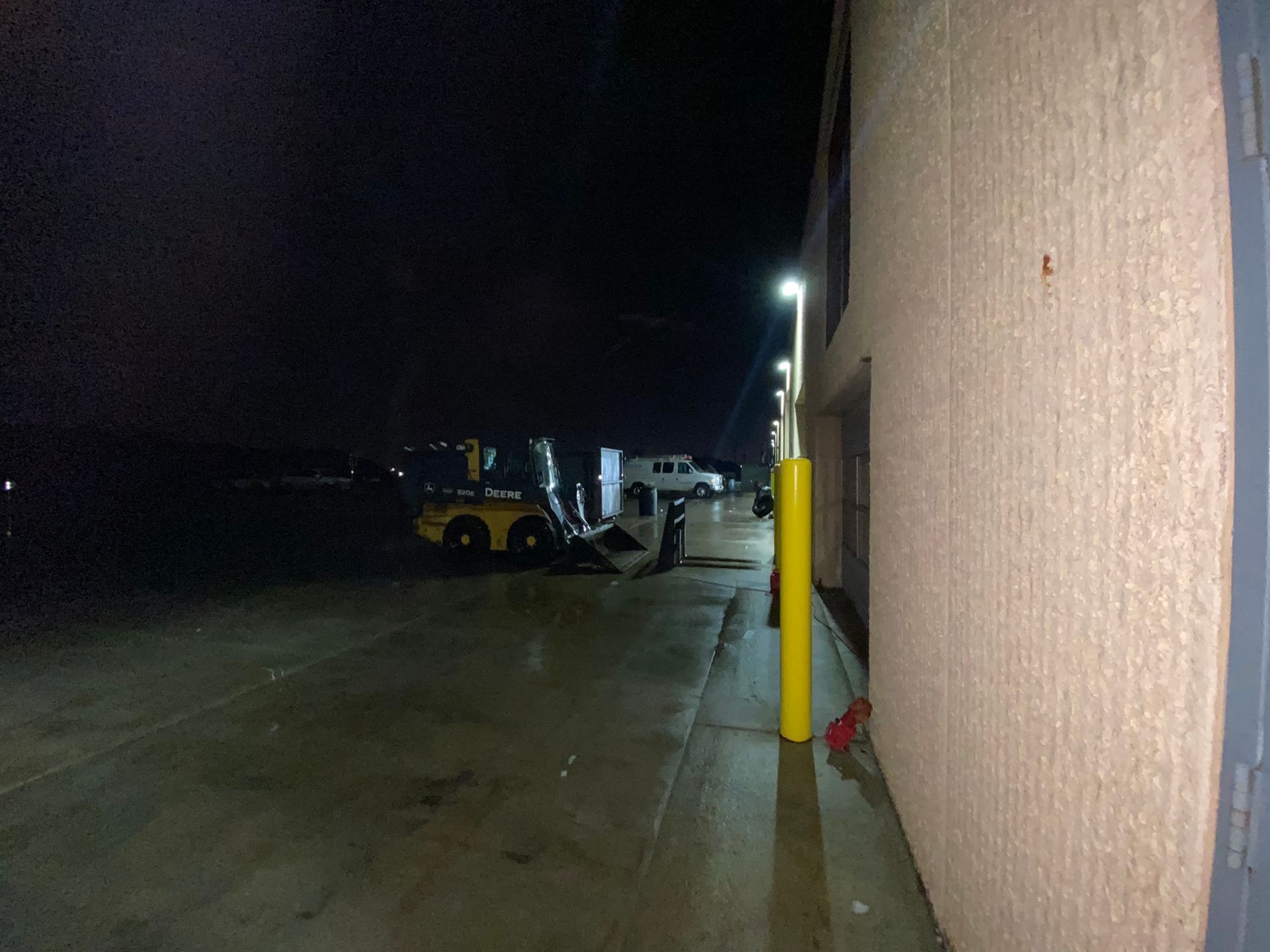 A dark exterior with a wet ground, yellow bollards, and machinery near a building.