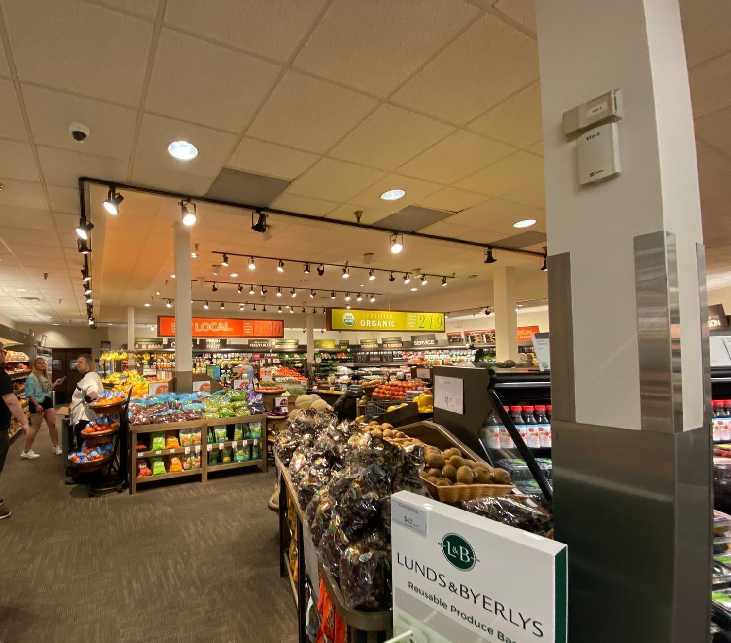 Grocery store interior with produce and shoppers; overhead lighting illuminates shelves and aisles.