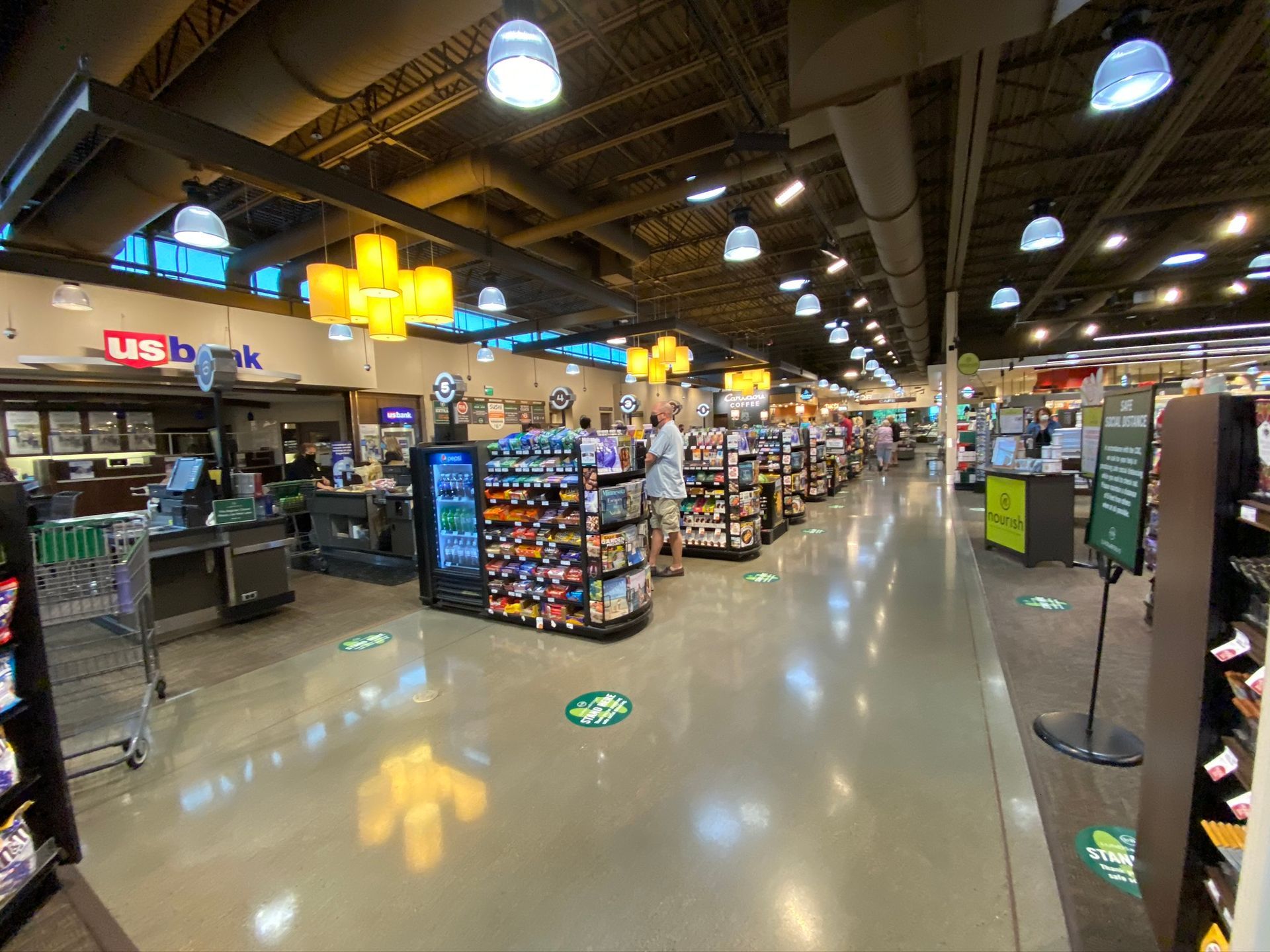 Interior view of a grocery store aisle with shoppers. Fluorescent lighting, concrete floor, and various product displays are visible.