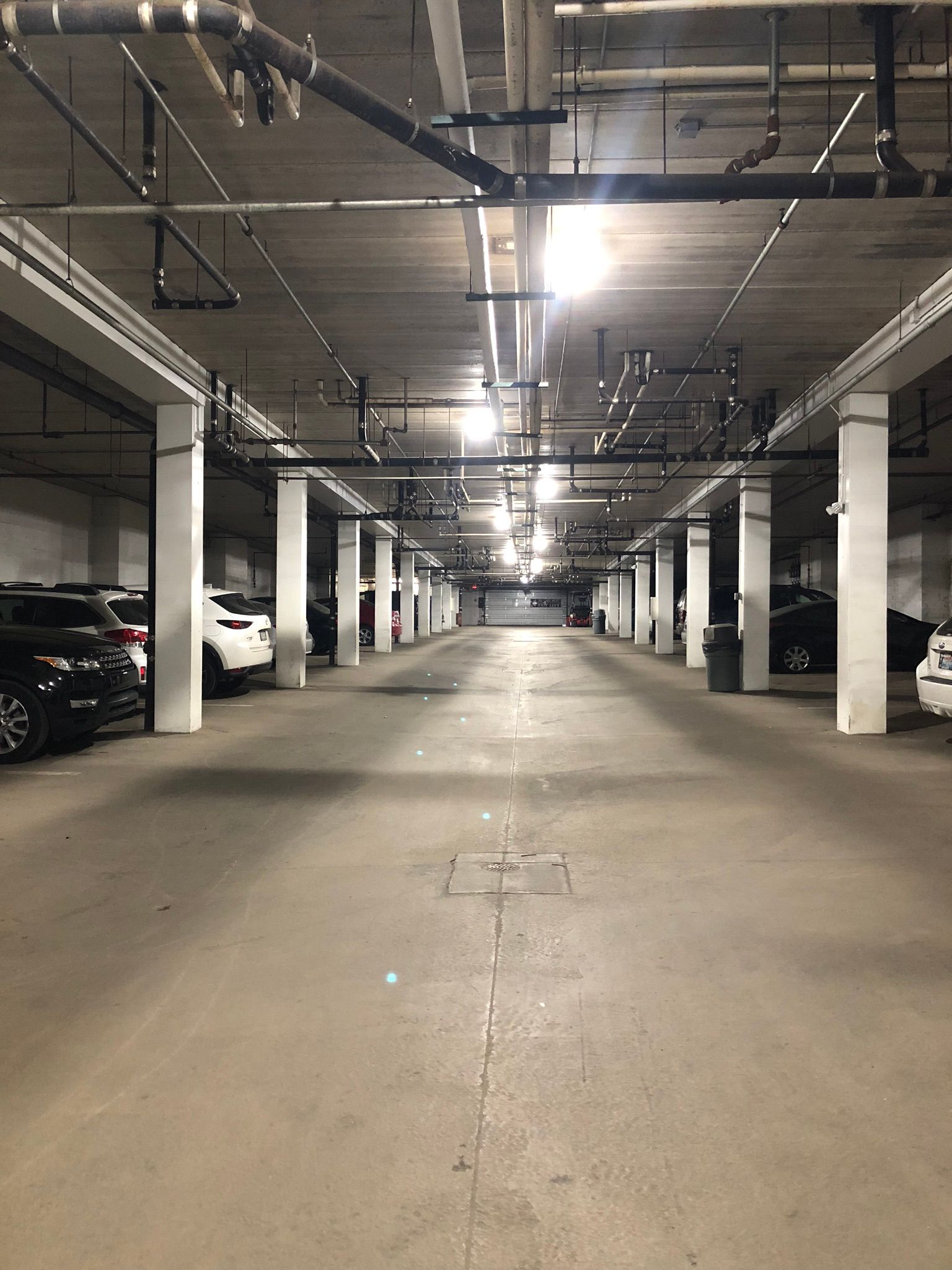 Underground parking garage with cars parked on either side, concrete floor, ceiling pipes, and bright lights.