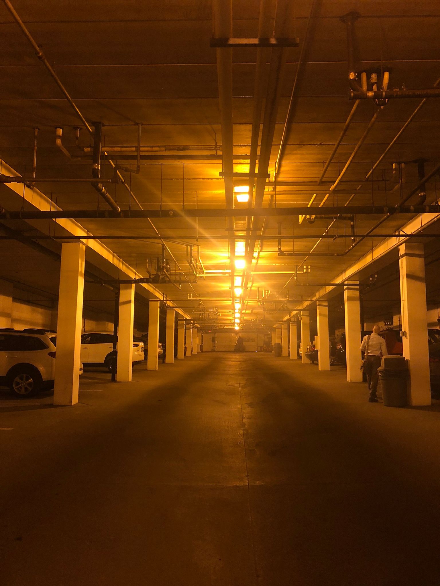 Indoor parking garage, long view. Rows of white columns, overhead lights illuminate the dark space. A person walks.