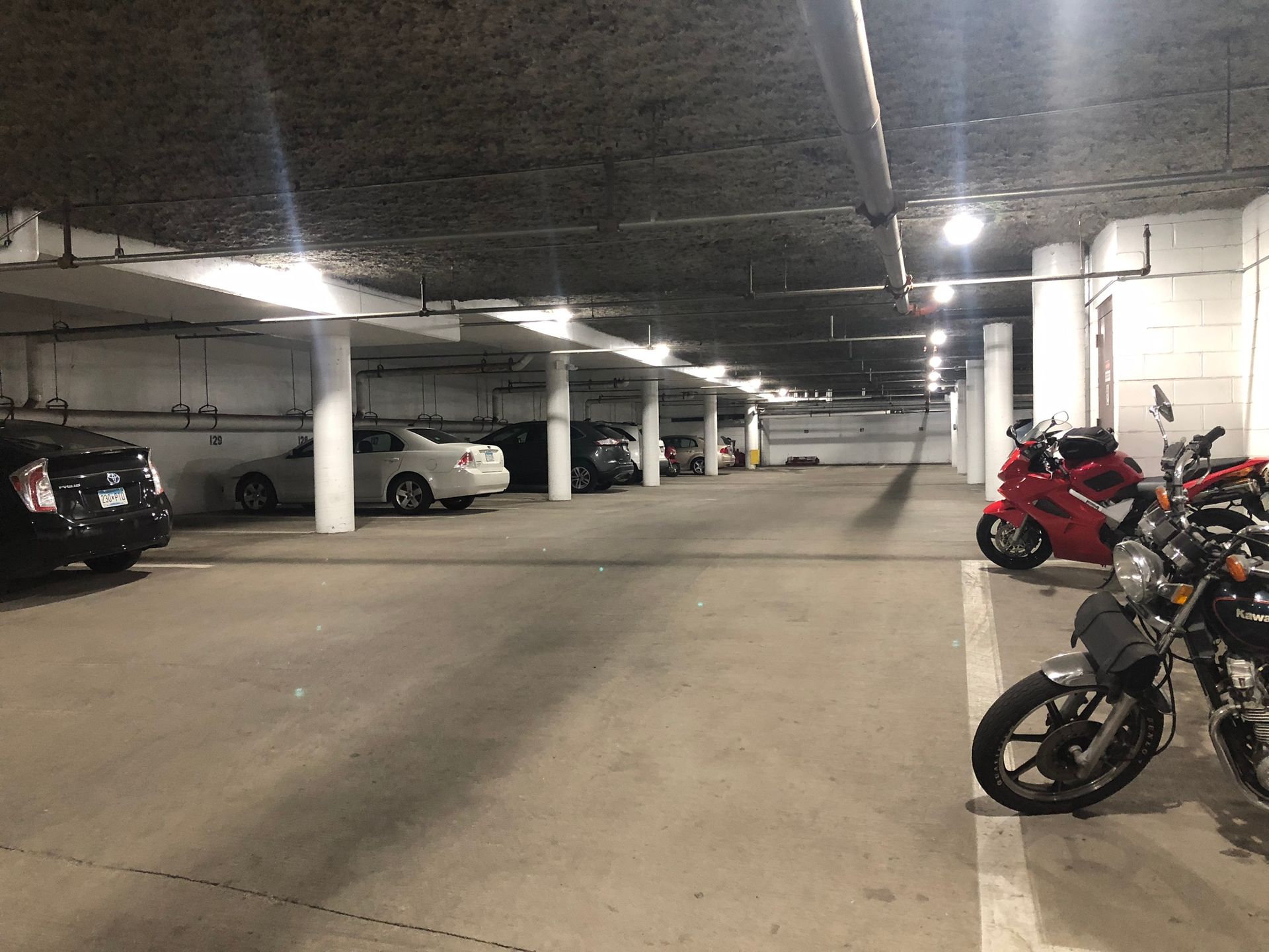 Underground parking garage with cars and motorcycles parked. Concrete columns and ceiling, overhead lighting.