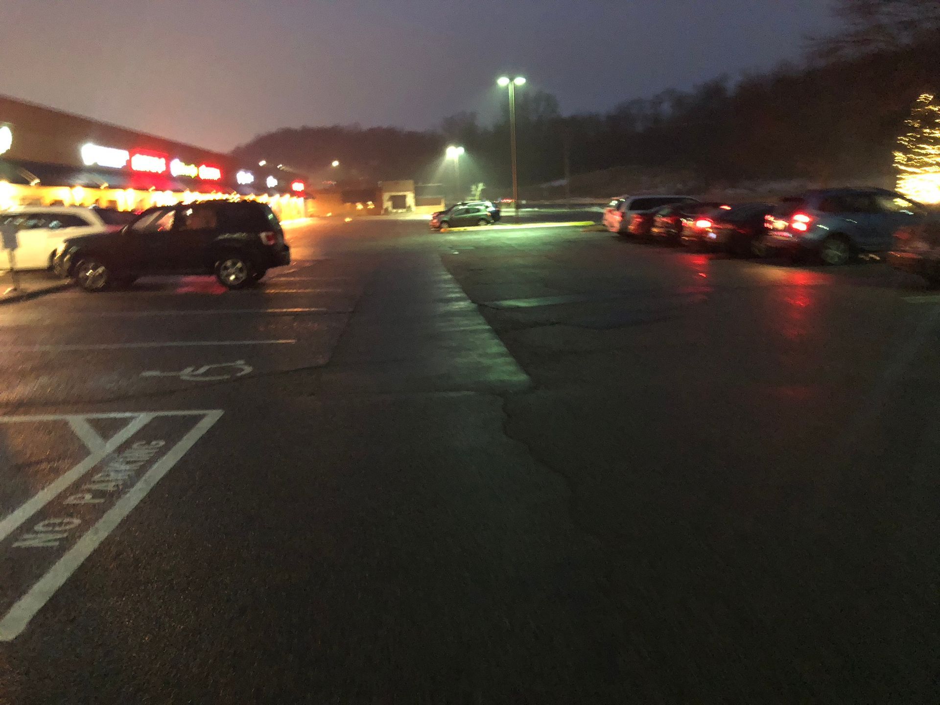 Dark parking lot at dusk, with cars parked near lit storefronts and streetlights.