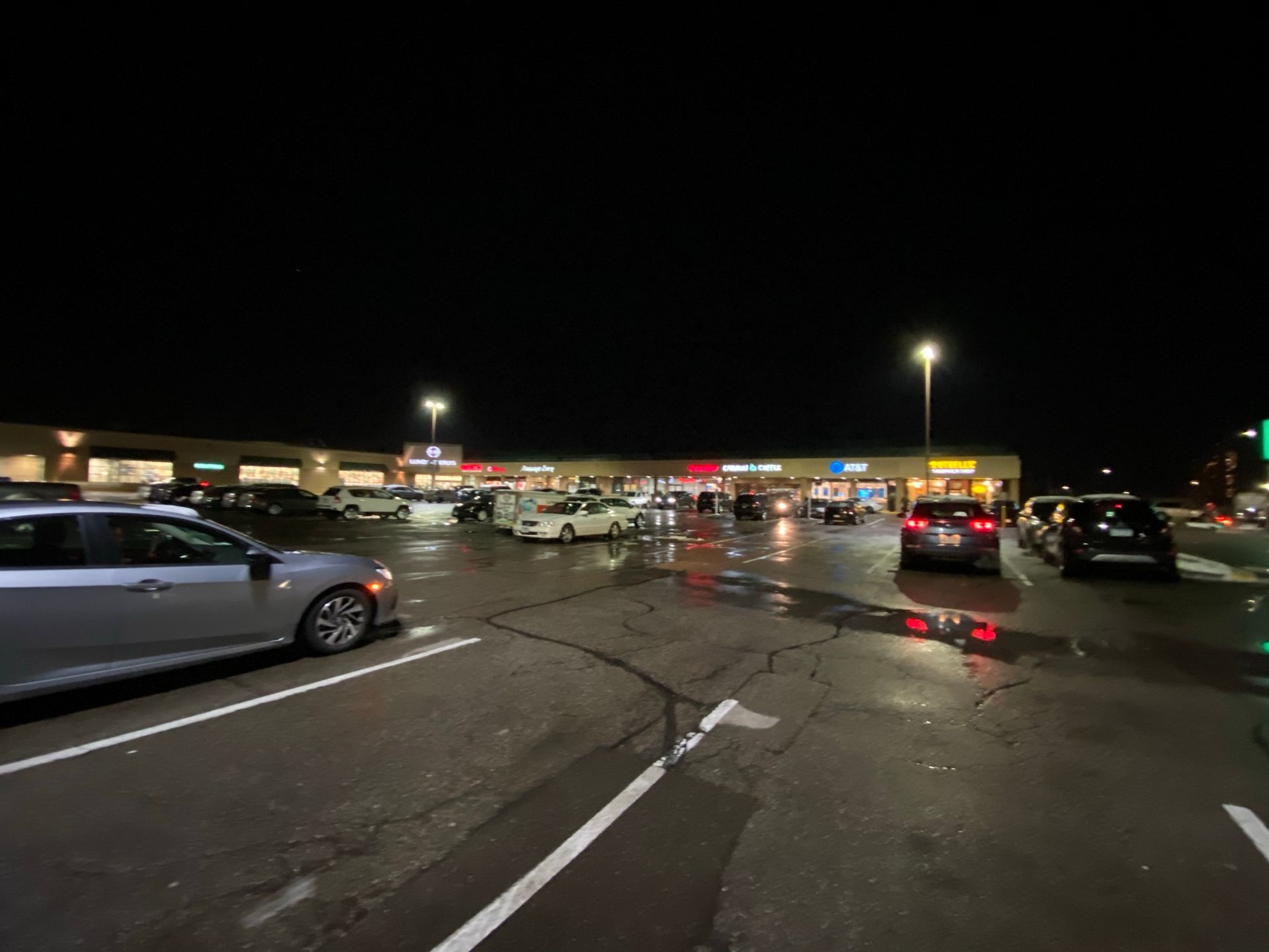 Nighttime shot of a strip mall parking lot with cars and storefronts under bright streetlights. Wet asphalt reflects lights.