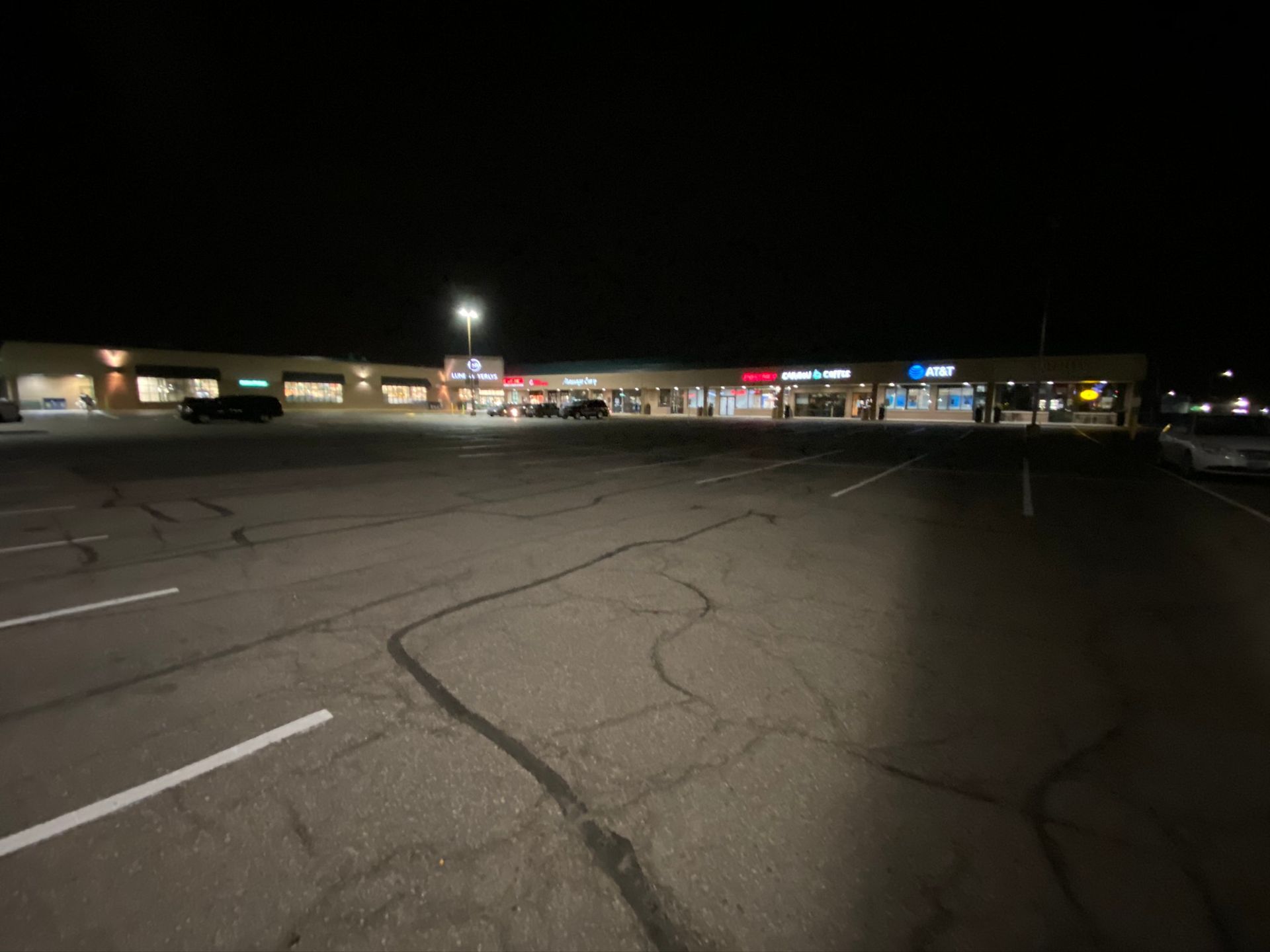 Empty parking lot in front of a strip mall at night with lights on.