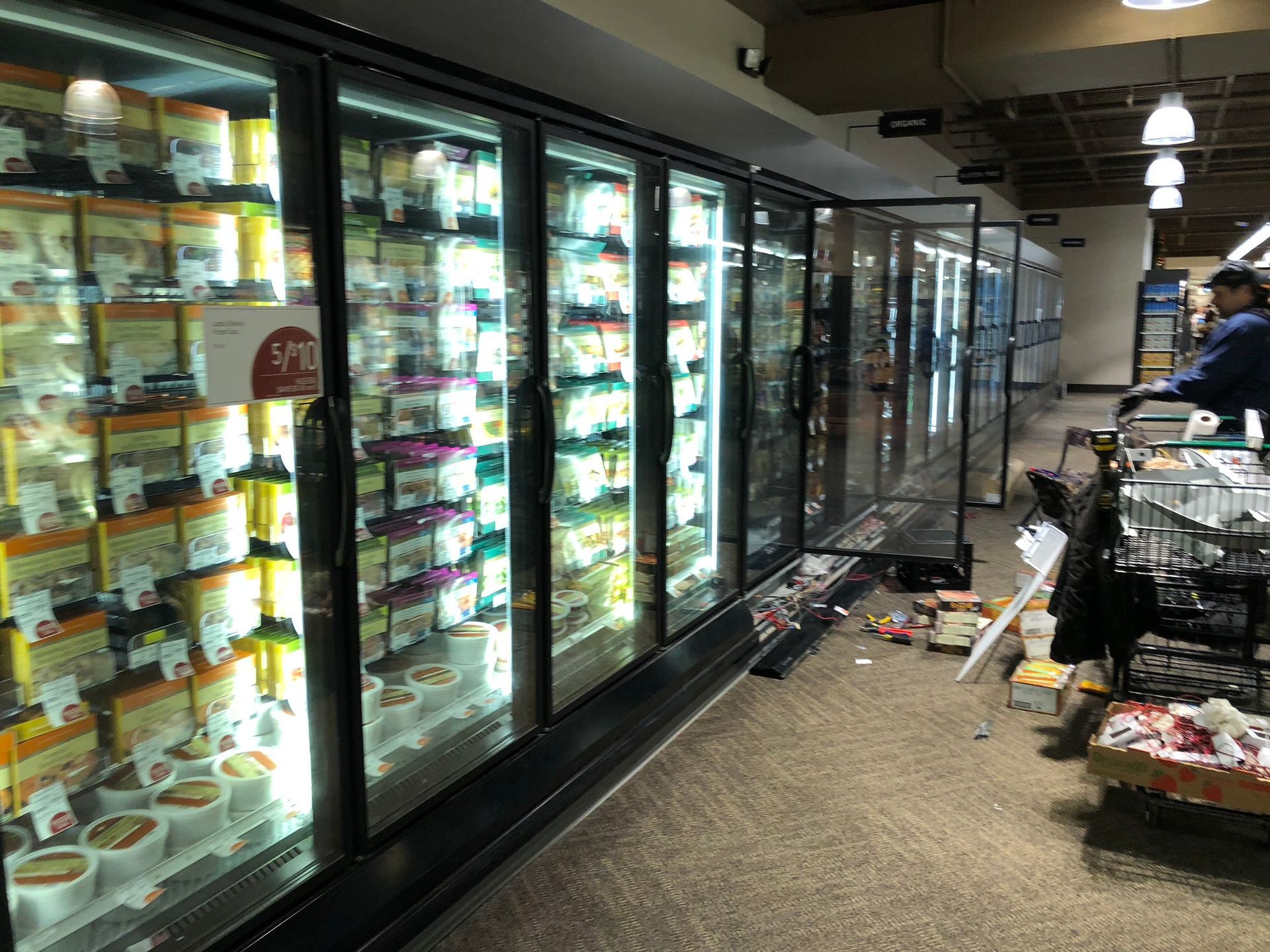 Refrigerated food display case in a grocery store, with open door. A person is nearby. Debris on the floor.