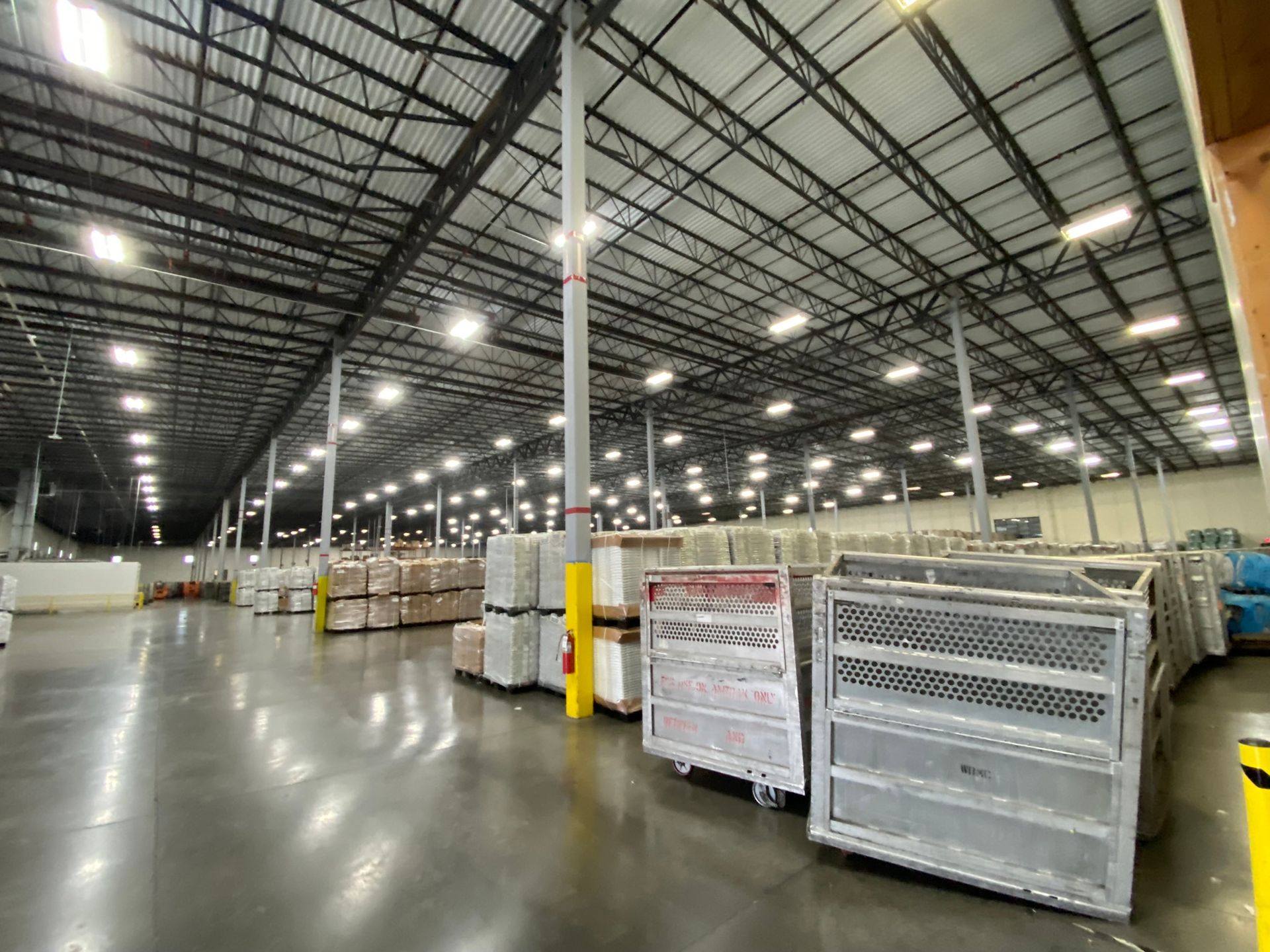 An expansive, brightly lit warehouse interior with stacks of pallets and large, wheeled metal sorting bins on the floor.