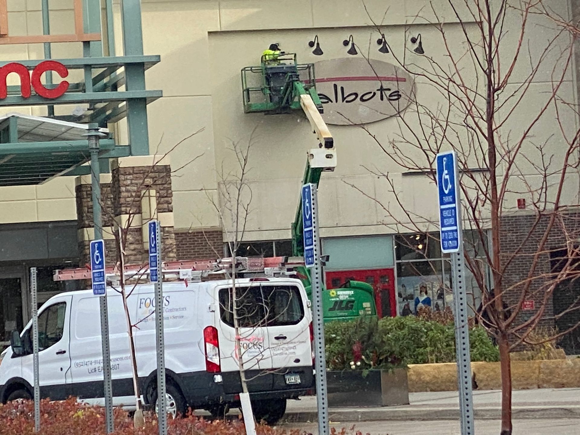 A worker in a bucket truck repairs the sign of a Talbots store near an AMC theater and a white service van.