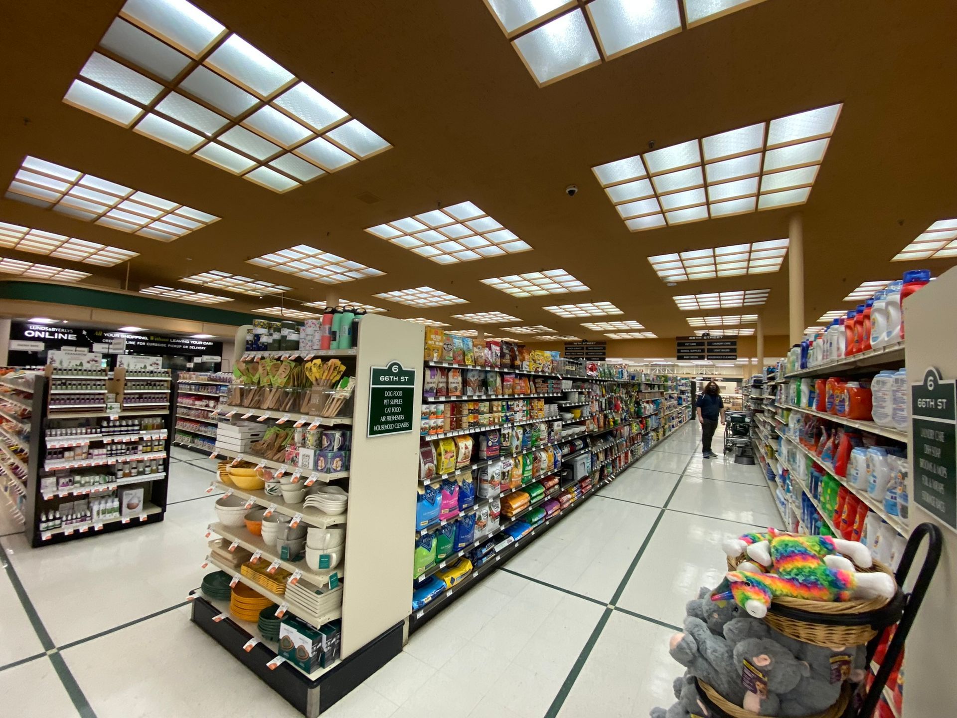 Grocery store interior with rows of shelves, a person shopping, and bright overhead lighting.