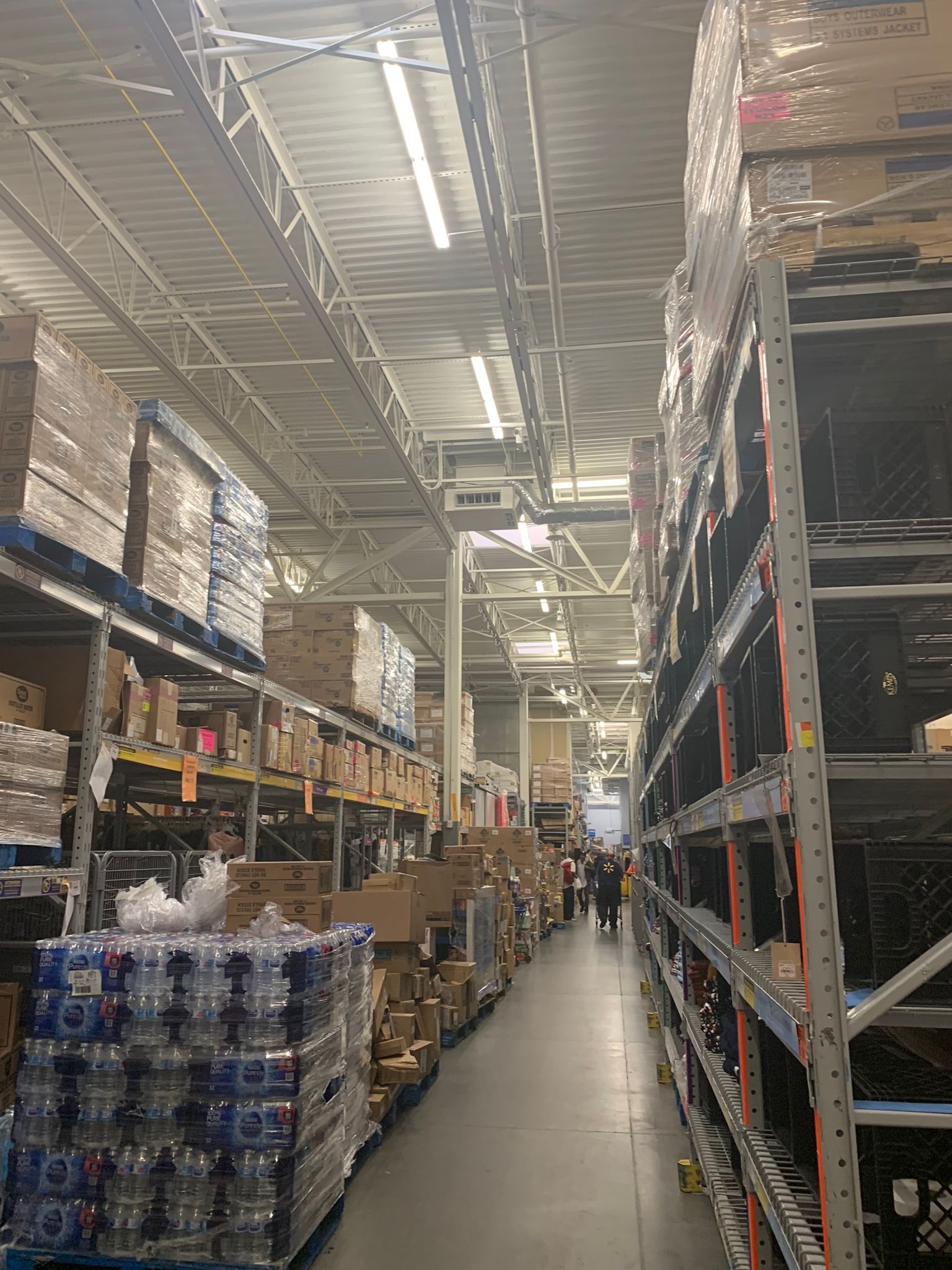 Warehouse aisle with boxes and shelves, overhead lighting, people visible in the distance.
