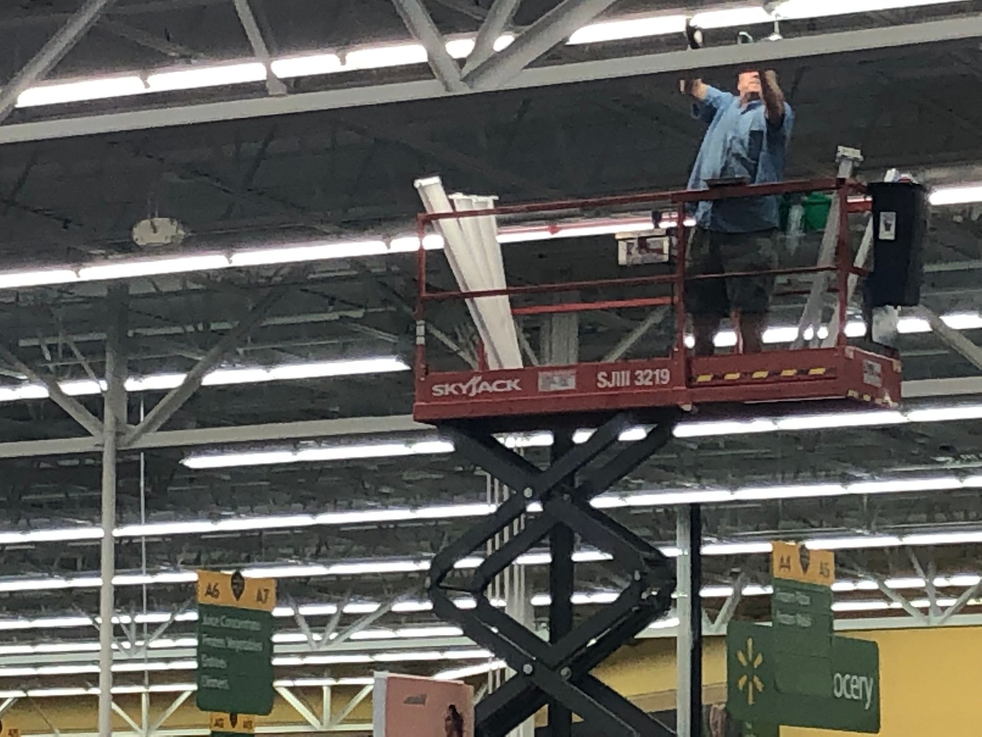 Man on a scissor lift installing ceiling fixtures in a large retail store with white support beams.